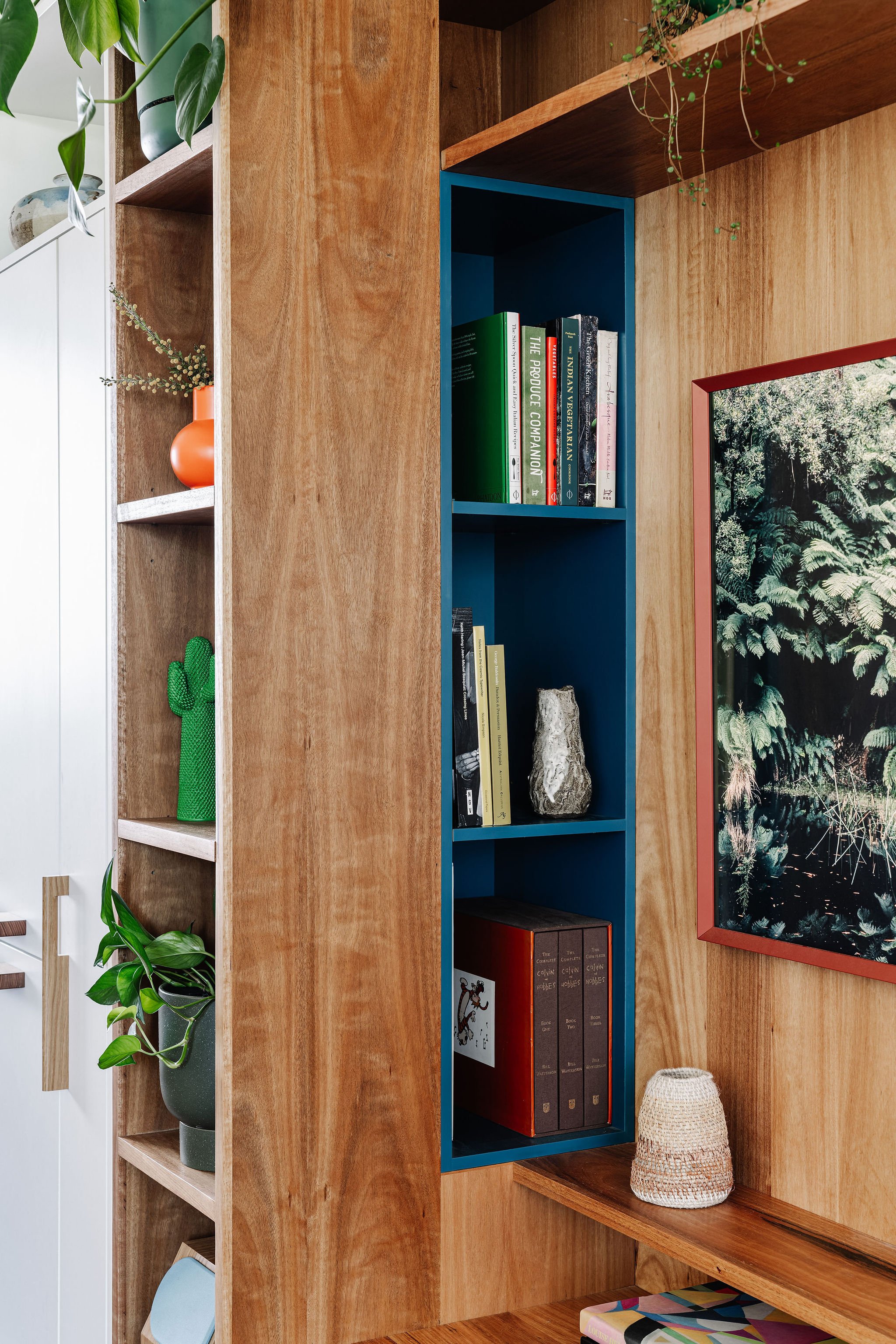 Wooden Bookshelf with books, plants, decorative vases, and a framed picture of a lush green forest.
