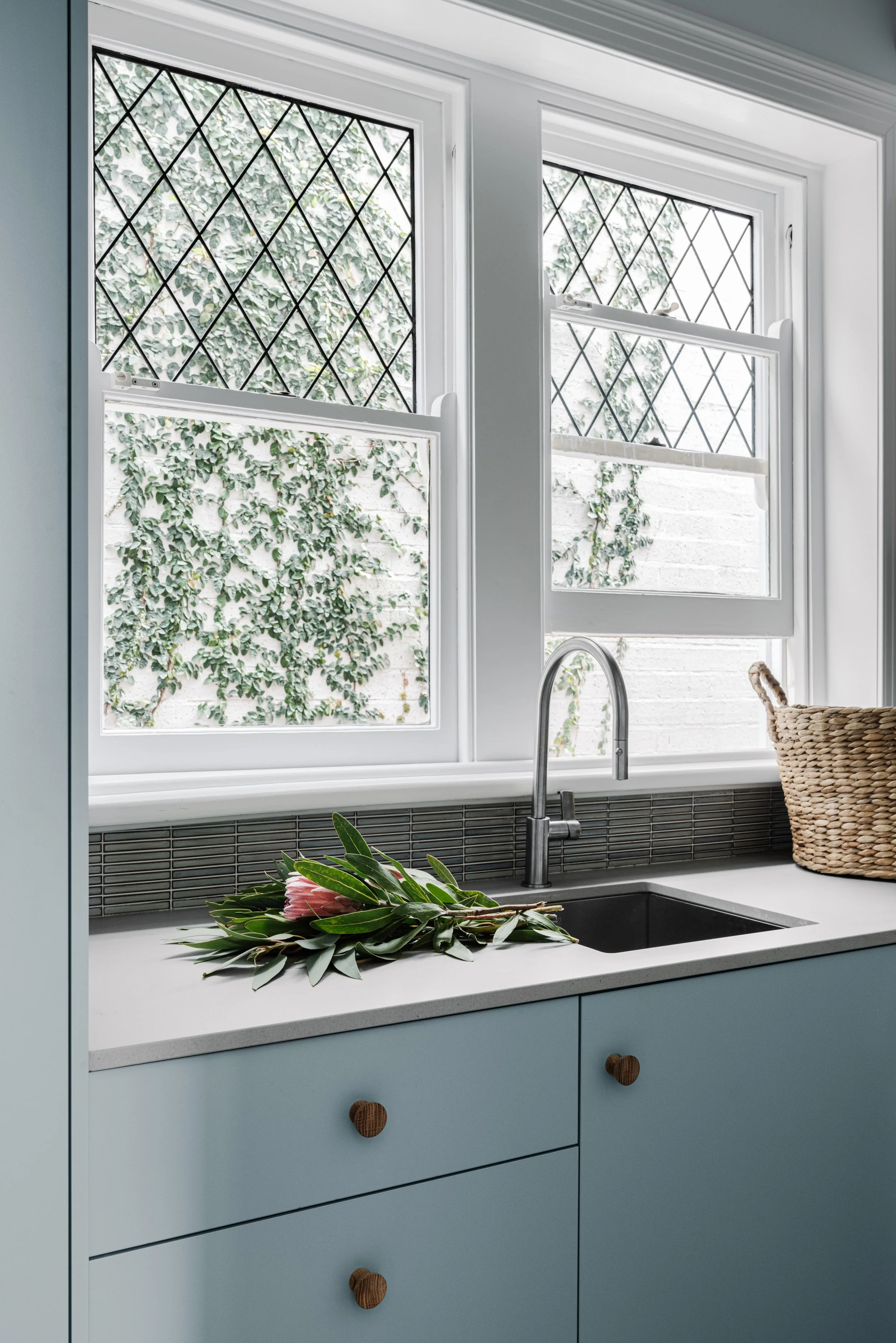 Kitchen window with black diamond-patterned security grille and a view of greenery outside. Inside, a white countertop with a sink, a bundle of flowers, and a woven basket.