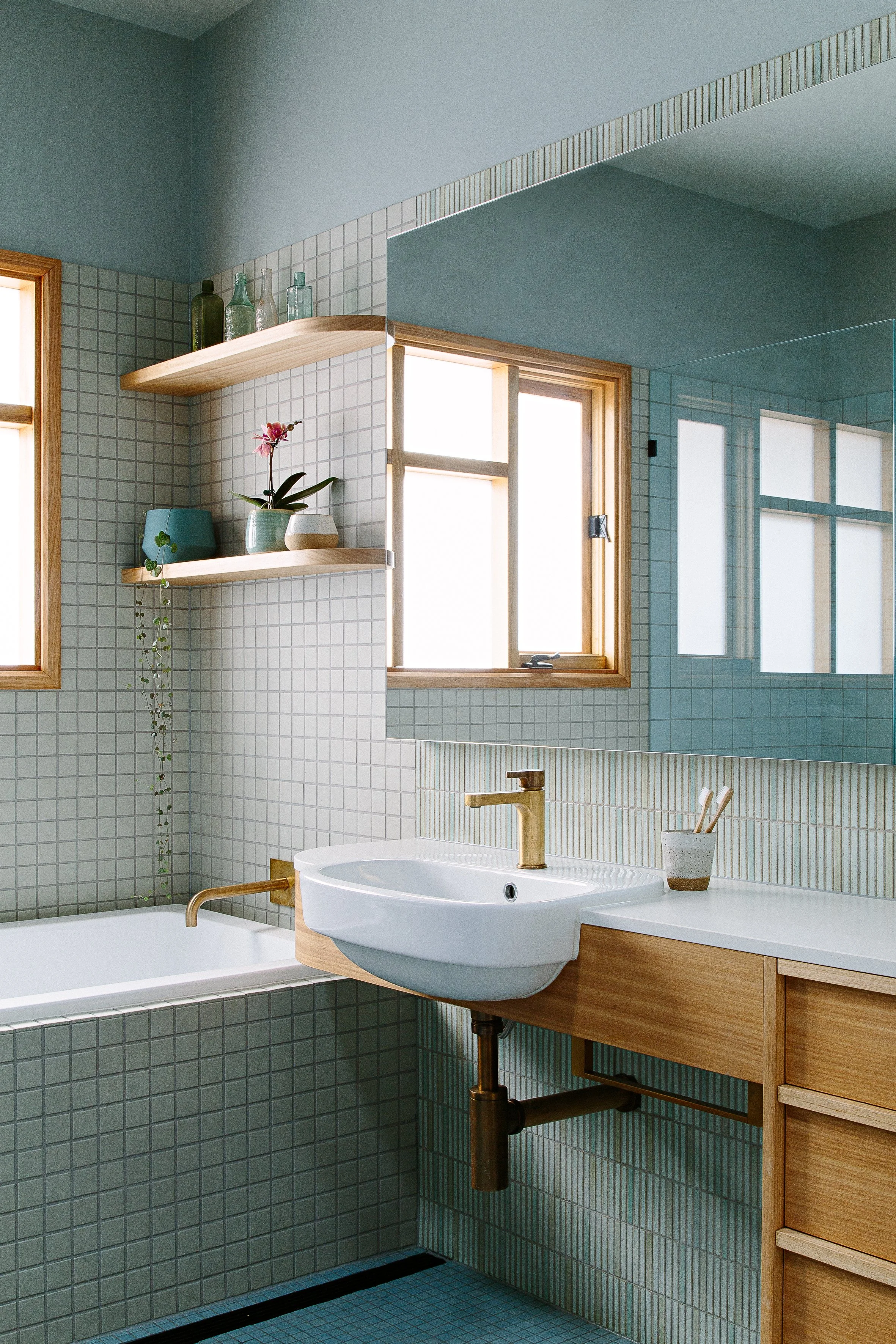 Bathroom with green square tiles, wooden window frames, a white sink with a gold faucet, and floating wooden shelves with decorative bottles and plants.