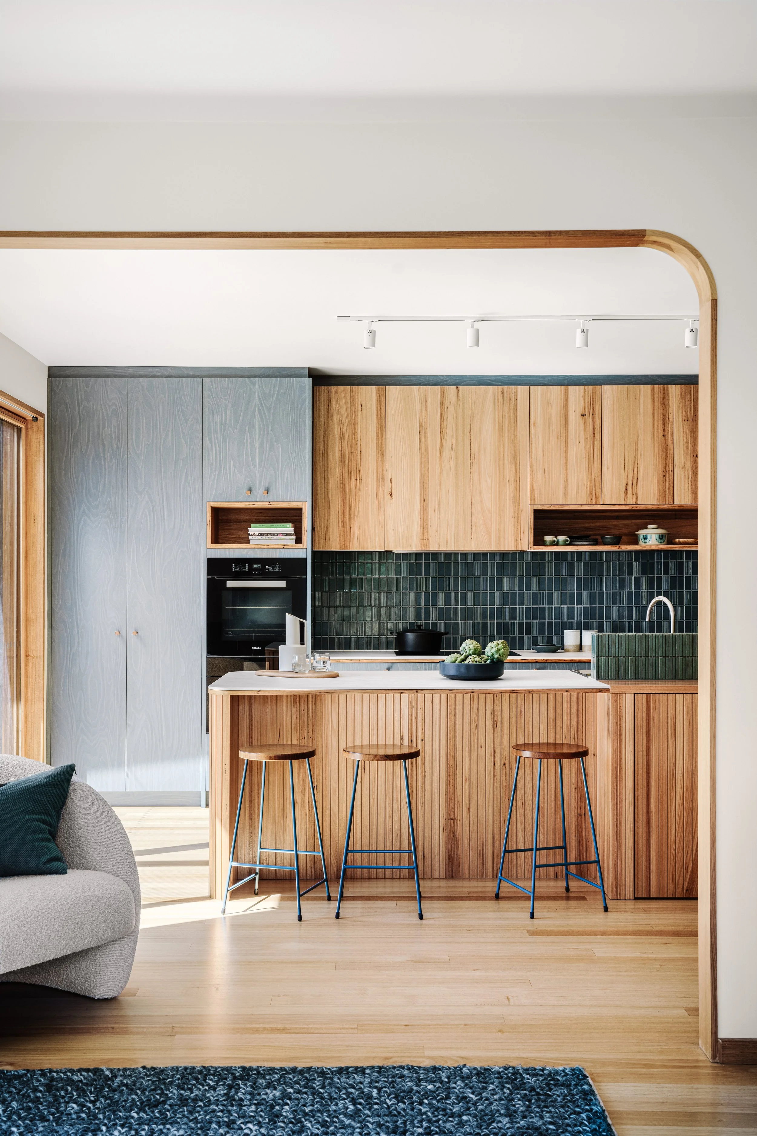 Open kitchen with wooden cabinetry, dark green tile backsplash, and a kitchen island with three stools. A bowl of artichokes is on the island, and there's a row of track lighting on the ceiling.