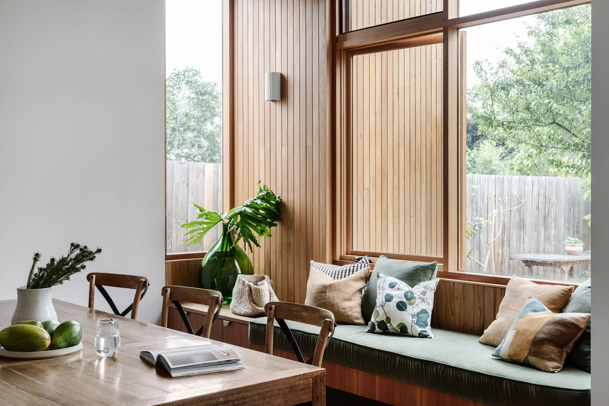 A cozy living room with natural wood paneling on the walls, large windows showing green trees outside, a wooden bench with cushions, a table with a plant in a green vase, and some mangoes and a glass of water.