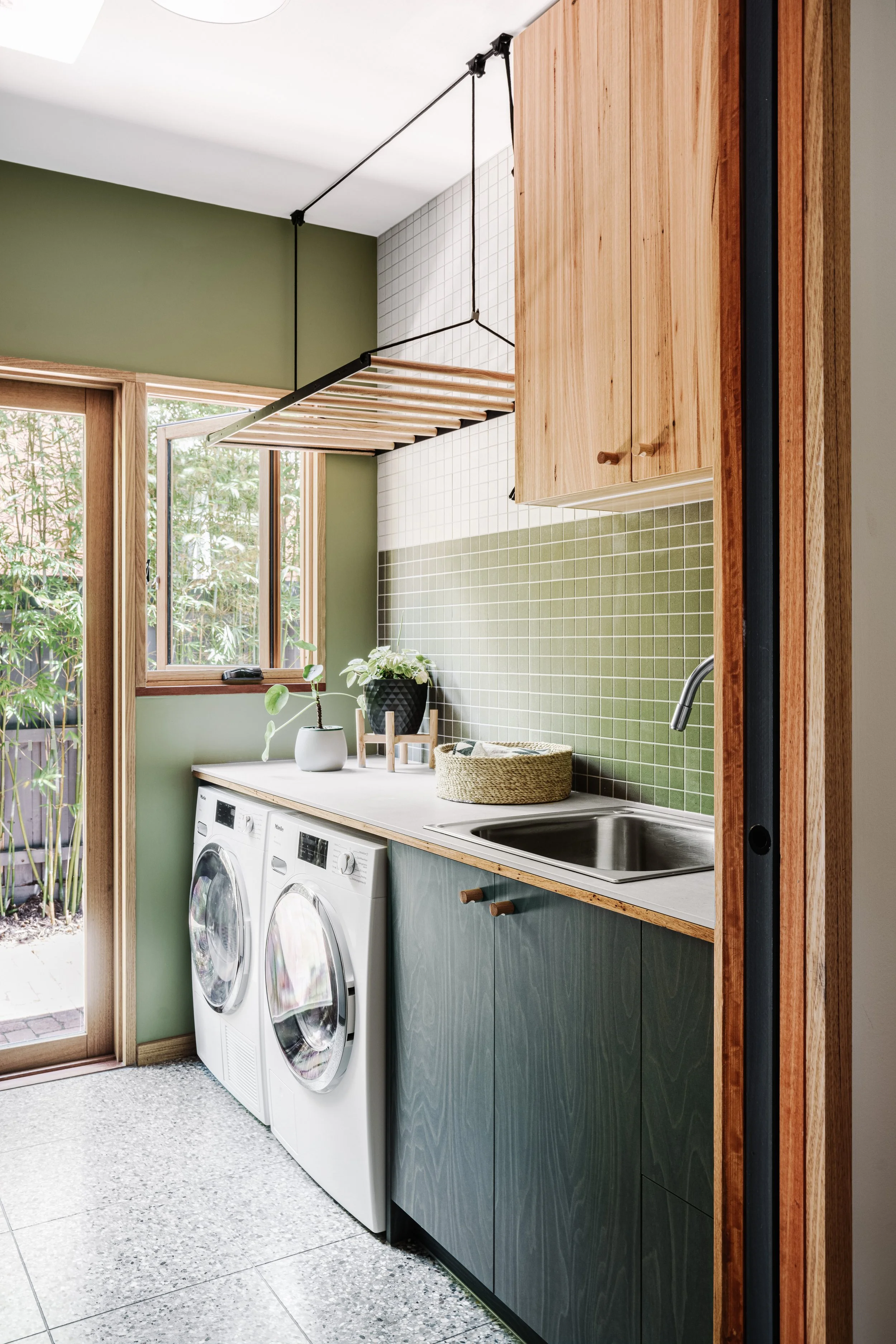 A laundry room with a washing machine and dryer beneath a white countertop. There are green cabinets with wooden handles above the counter, and a green tiled backsplash. A small plant in a black pot and a wicker basket are on the countertop. The room has a door with glass panels, revealing outside greenery, and a window letting in natural light.
