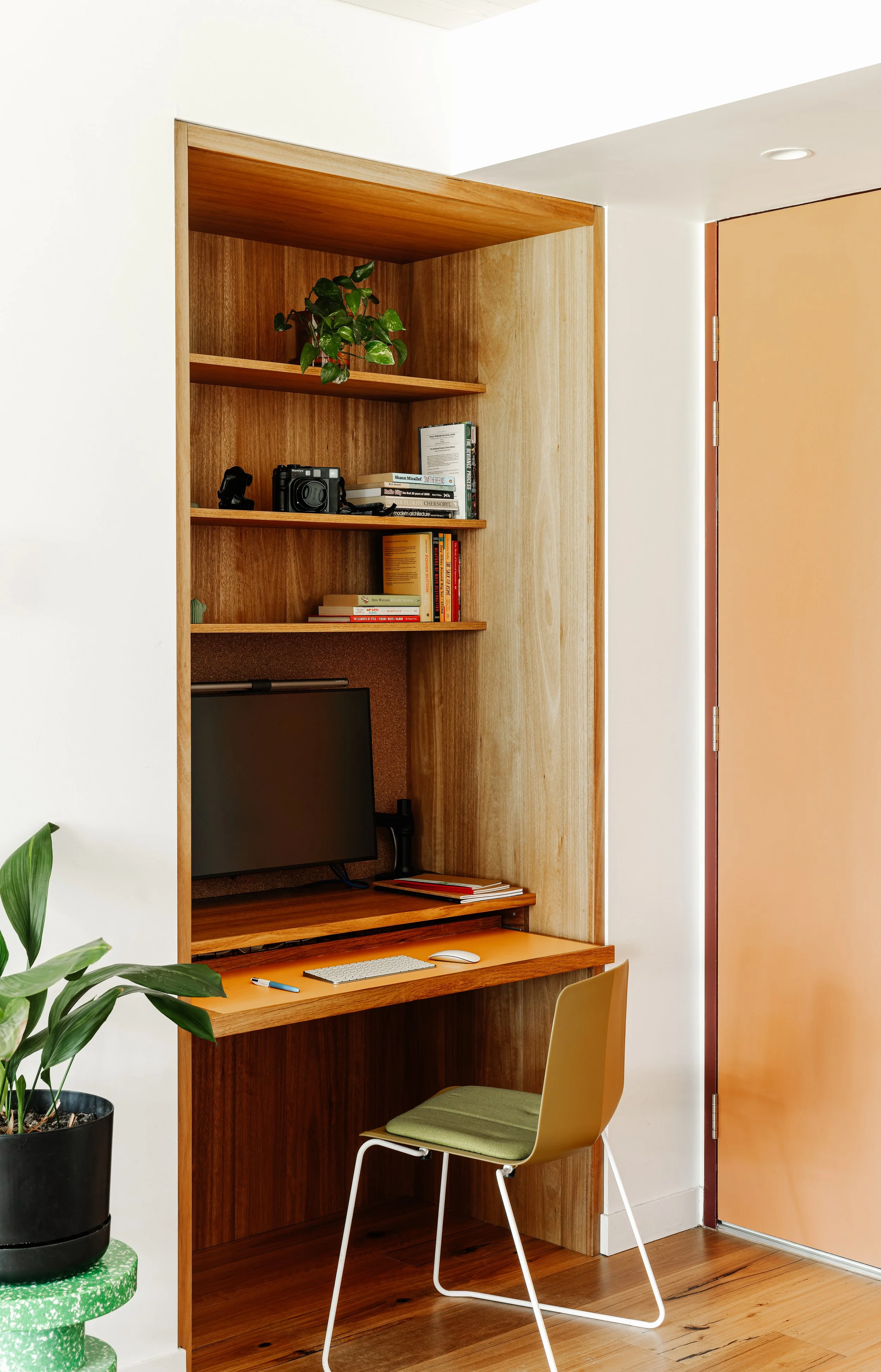 Home office workspace with wooden desk, yellow chair, computer monitor, bookshelf with books and plants, and a pink door.