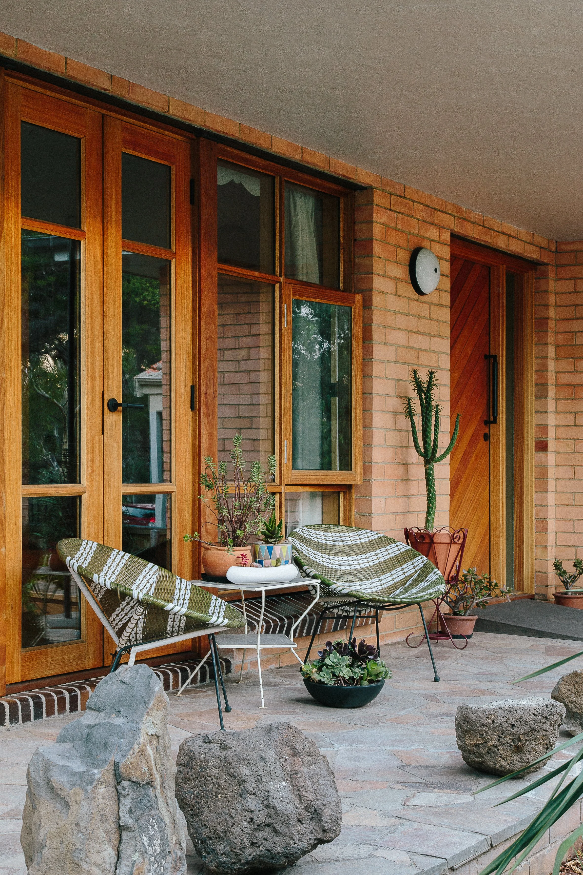 A cozy patio with wooden framed glass doors, potted succulents, rocking chaise lounges with green and white striped cushions, large rocks, and various potted plants on a stone tiled floor.