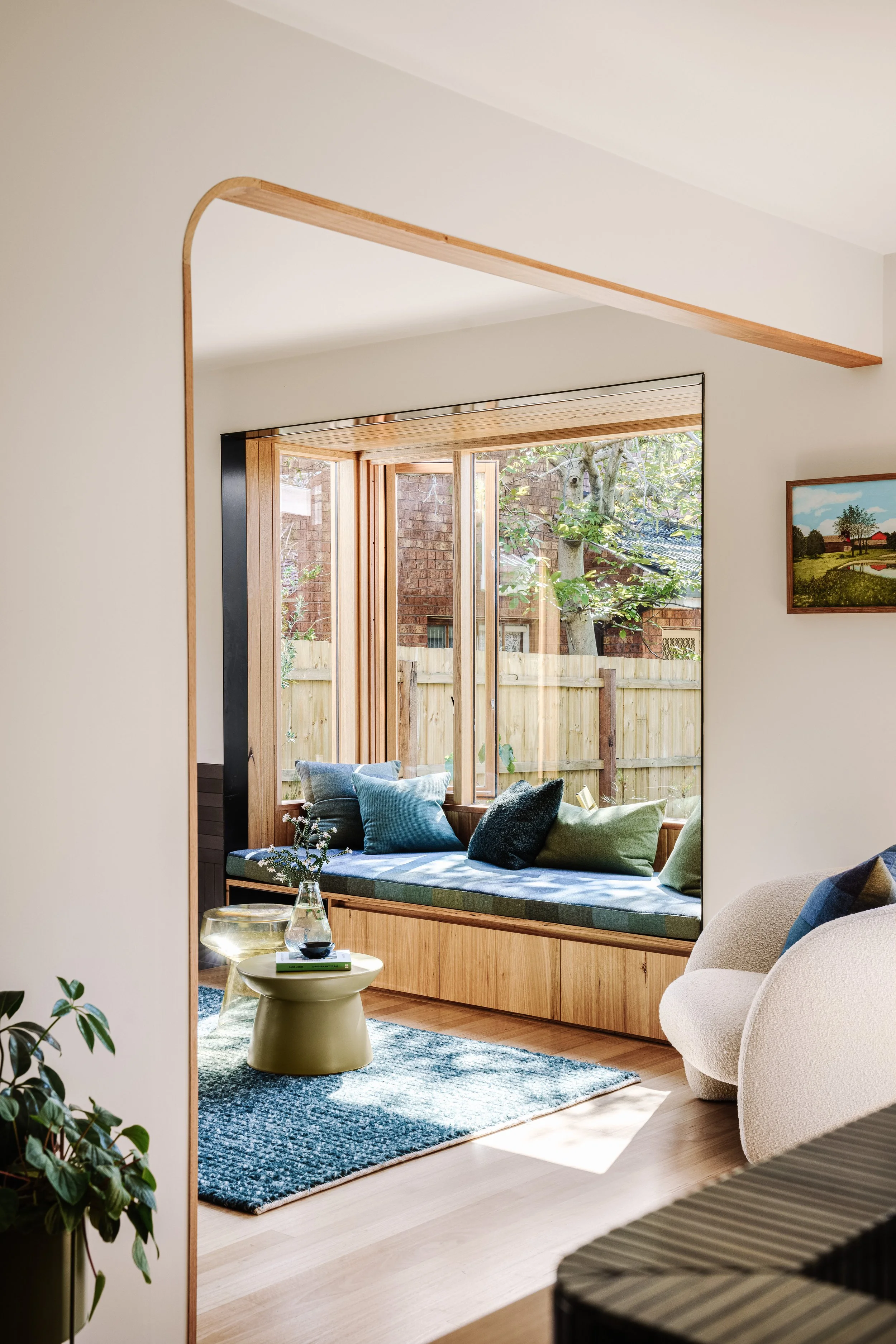 Living room with large window seat with cushions, framed by wooden trim, adjacent to a garden outside. A small round table with a glass vase and plant is in front of the window, with a beige armchair and framed picture on the wall inside.