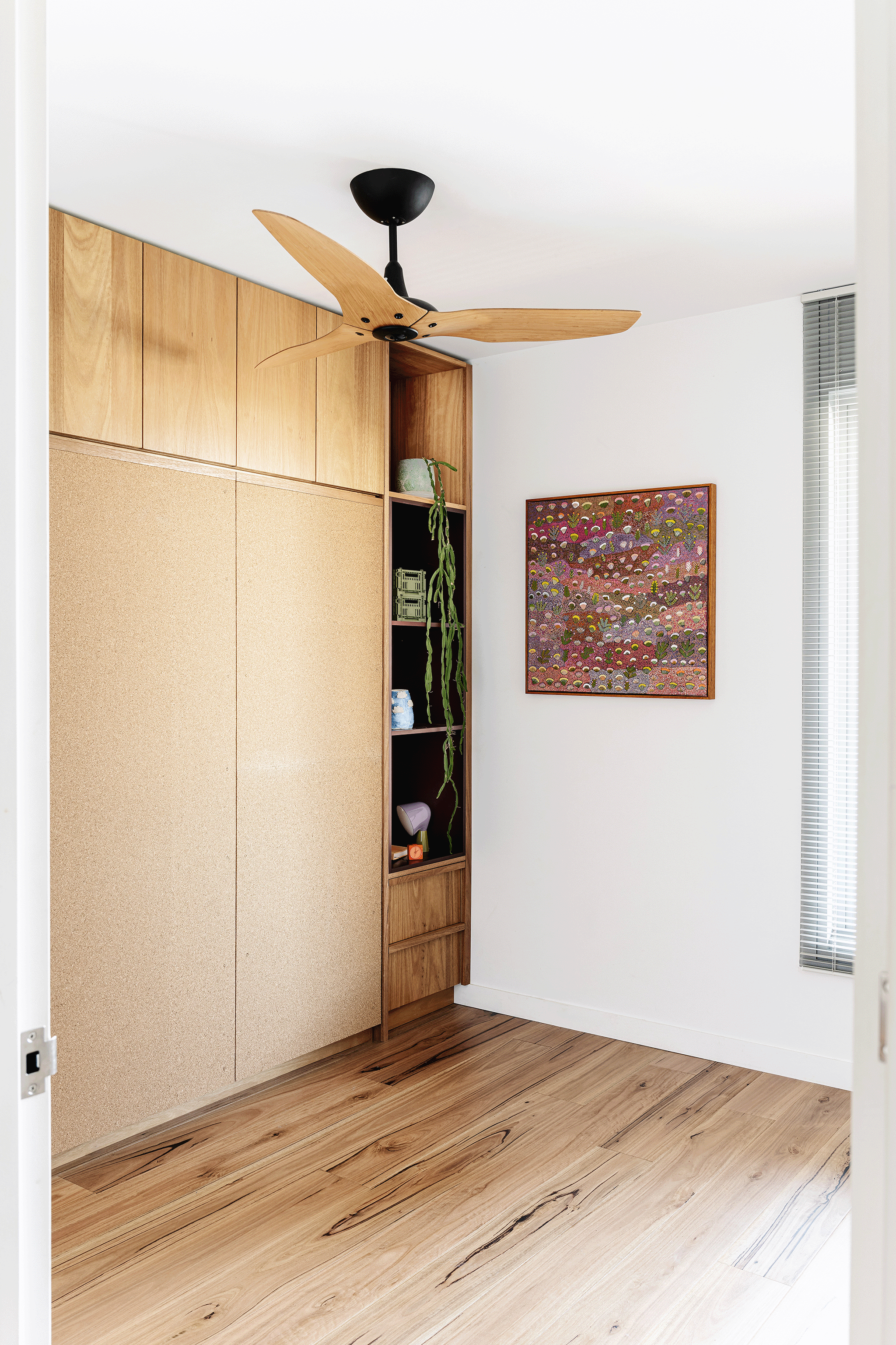 A corner of a room with a ceiling fan, a wooden cabinet with shelves, a framed colorful artwork on the wall, and a window with blinds.