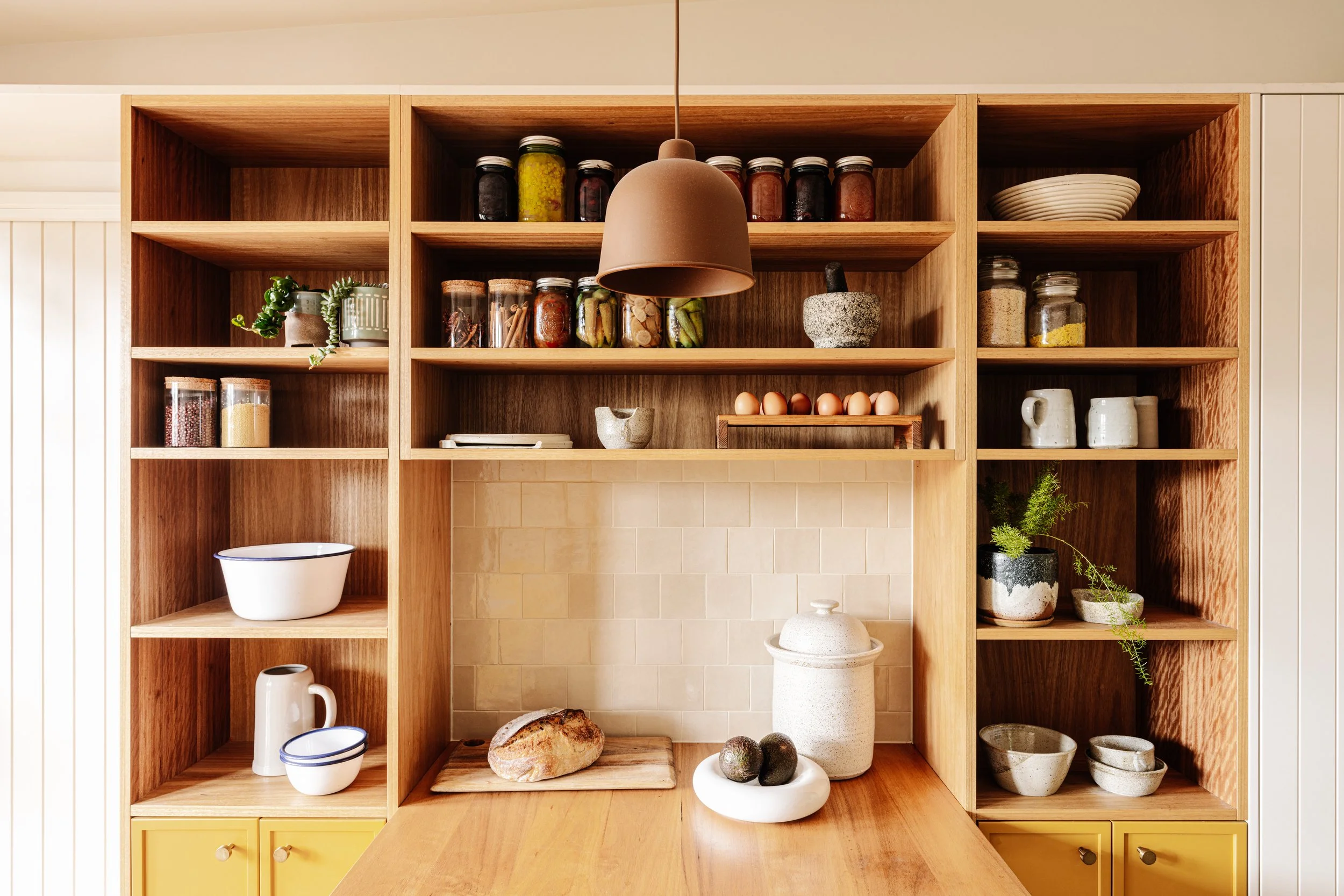 Open wooden kitchen shelving with jars of spices and preserves, bowls, a potted succulent, eggs, a loaf of bread, a large stone mortar and pestle, ceramic vases, and a yellow cabinet with drawers.