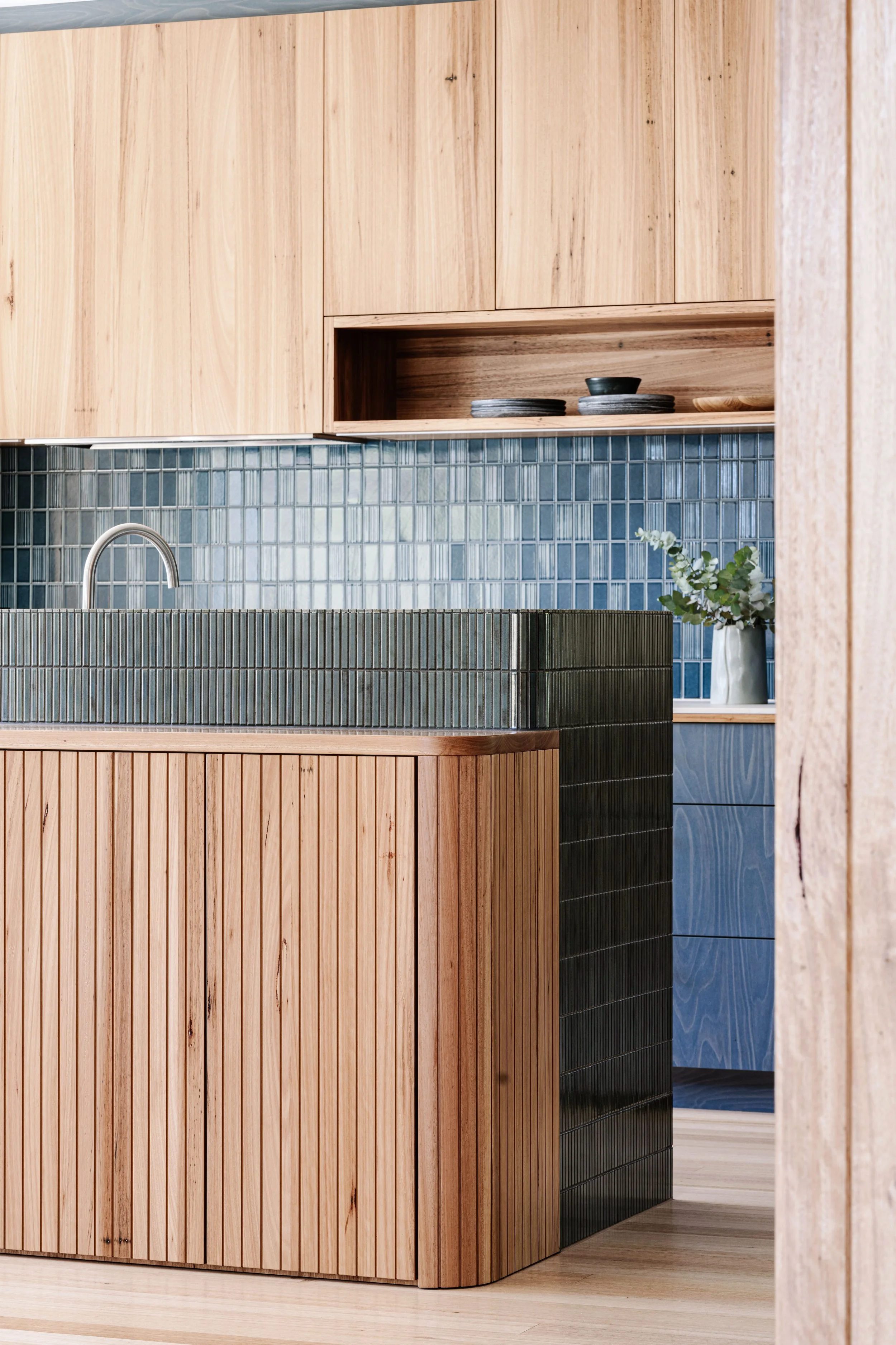 Wooden kitchen cabinet with open shelf displaying plates and a bowl, blue tile backsplash, a potted plant, and a sink with a curved faucet.