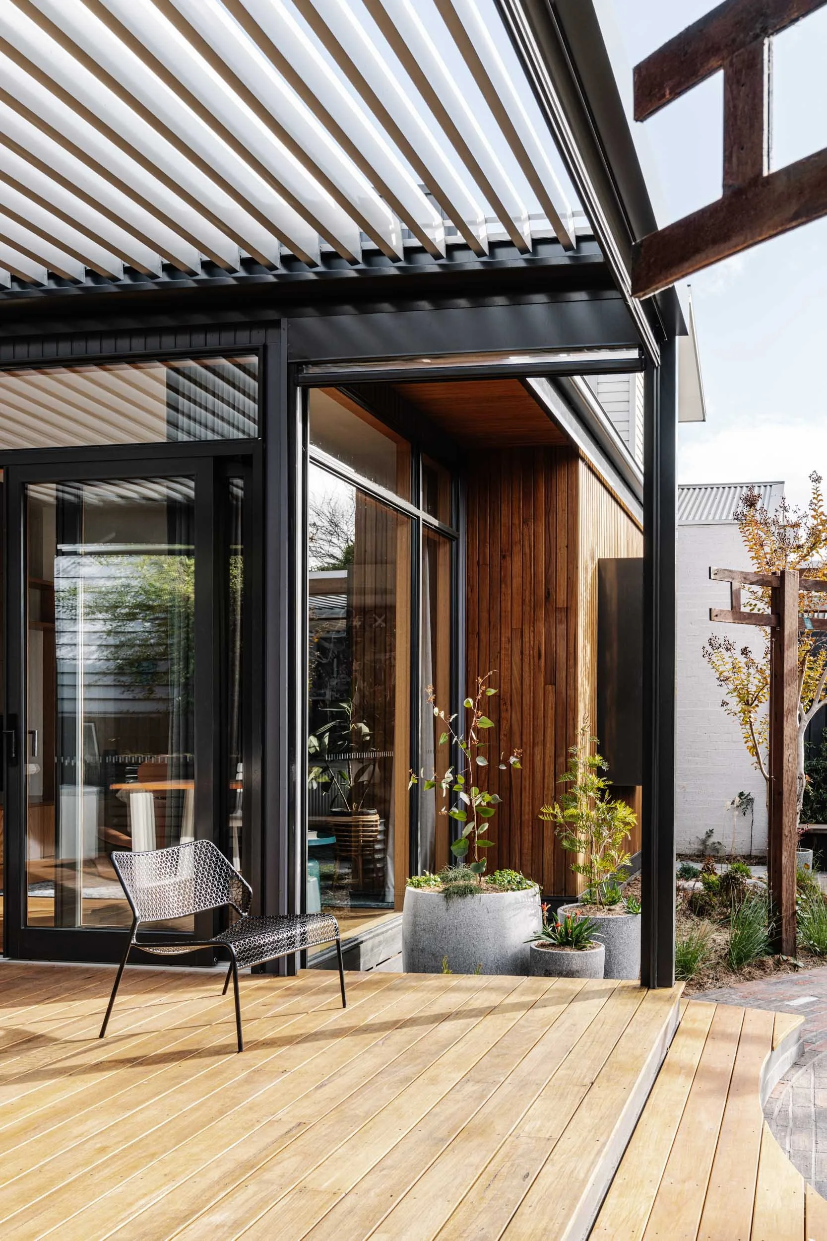 Modern house with large glass windows, wooden exterior, and an outdoor deck with potted plants and a black metal chair.