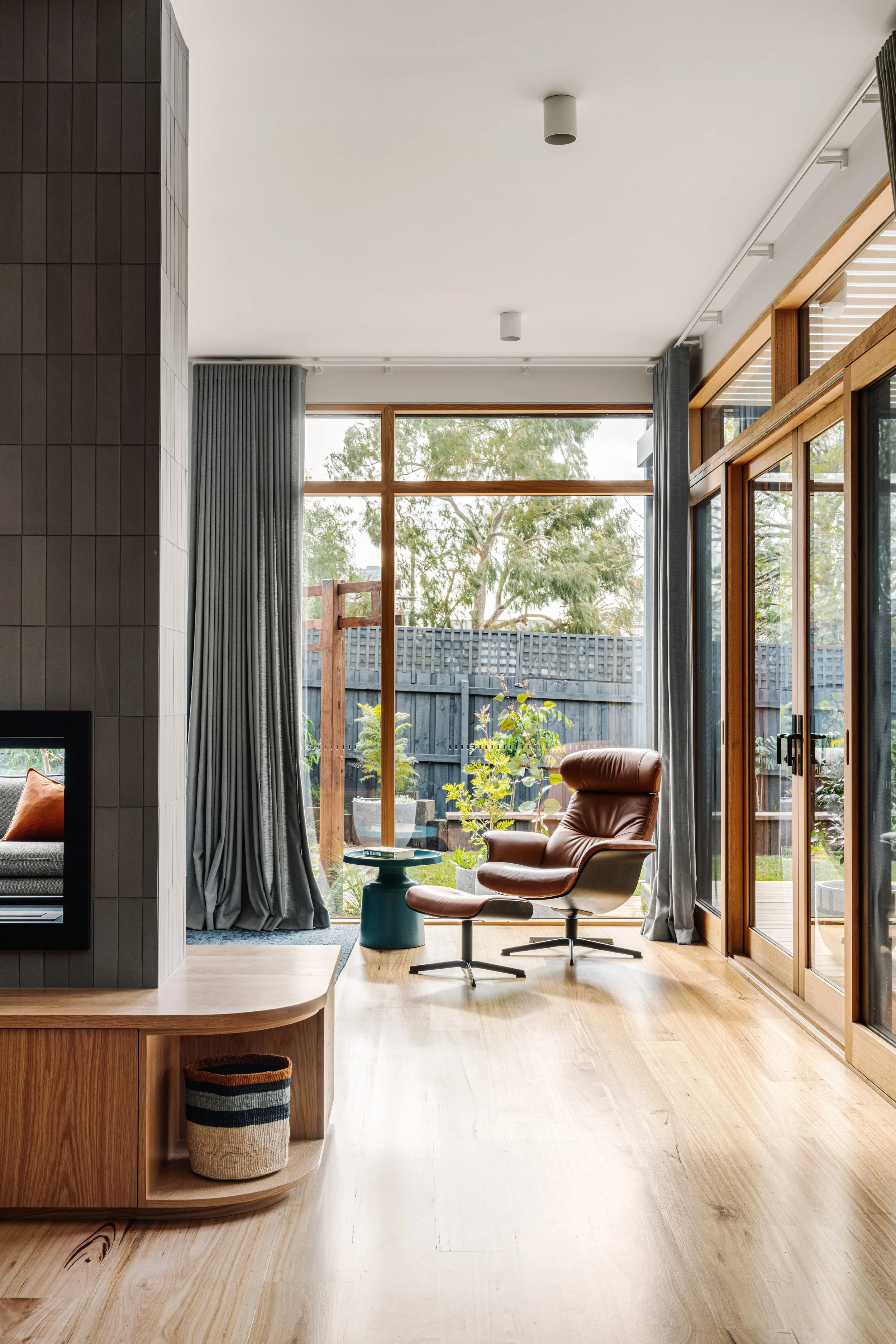 A modern interior living room with large floor-to-ceiling windows, wooden accents, and a cozy seating area with an orange chair and small side table, overlooking a backyard with plants and a wooden fence.