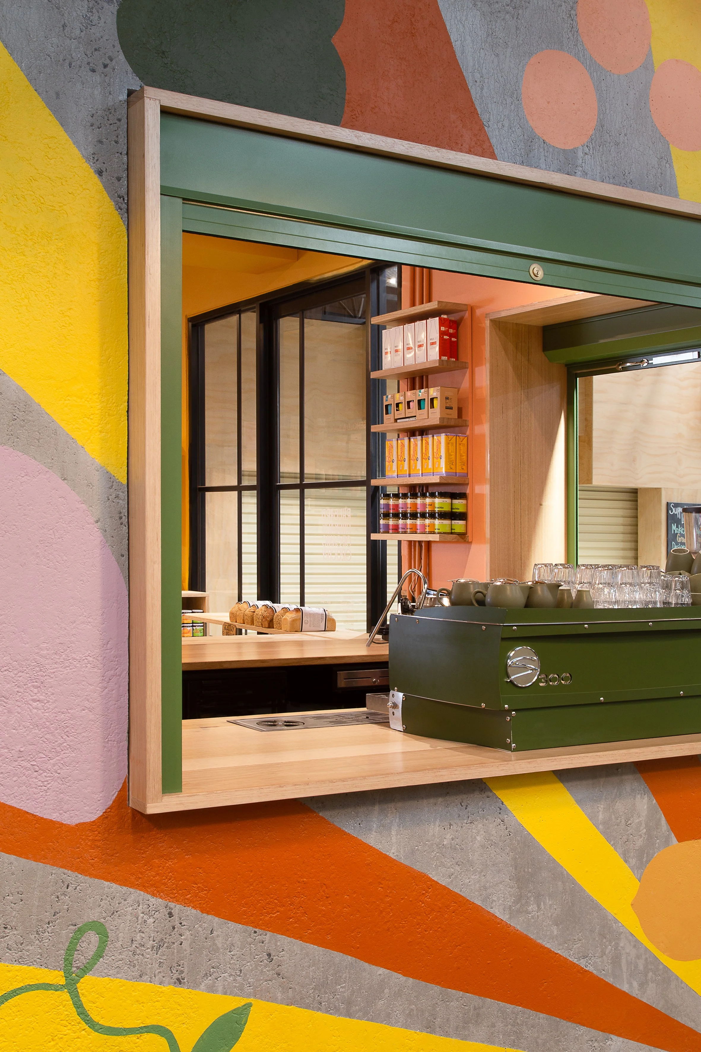 Colorful interior with mural wall art, a wooden counter with baked goods, shelves with boxes and jars, a large green commercial kitchen appliance, and large windows with black frames.
