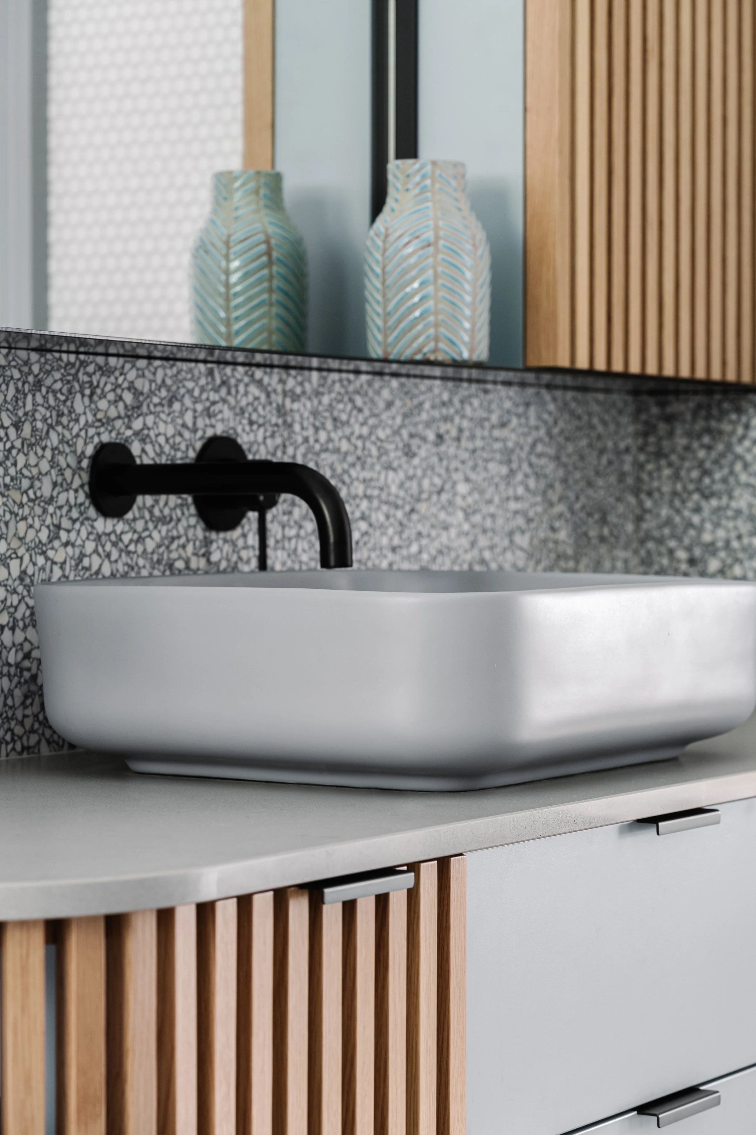 A bathroom with a terrazzo countertop, a white vessel sink, and a black faucet. There are decorative vases with a chevron pattern in the reflection of a mirror, and a wooden slatted cabinet below the countertop.