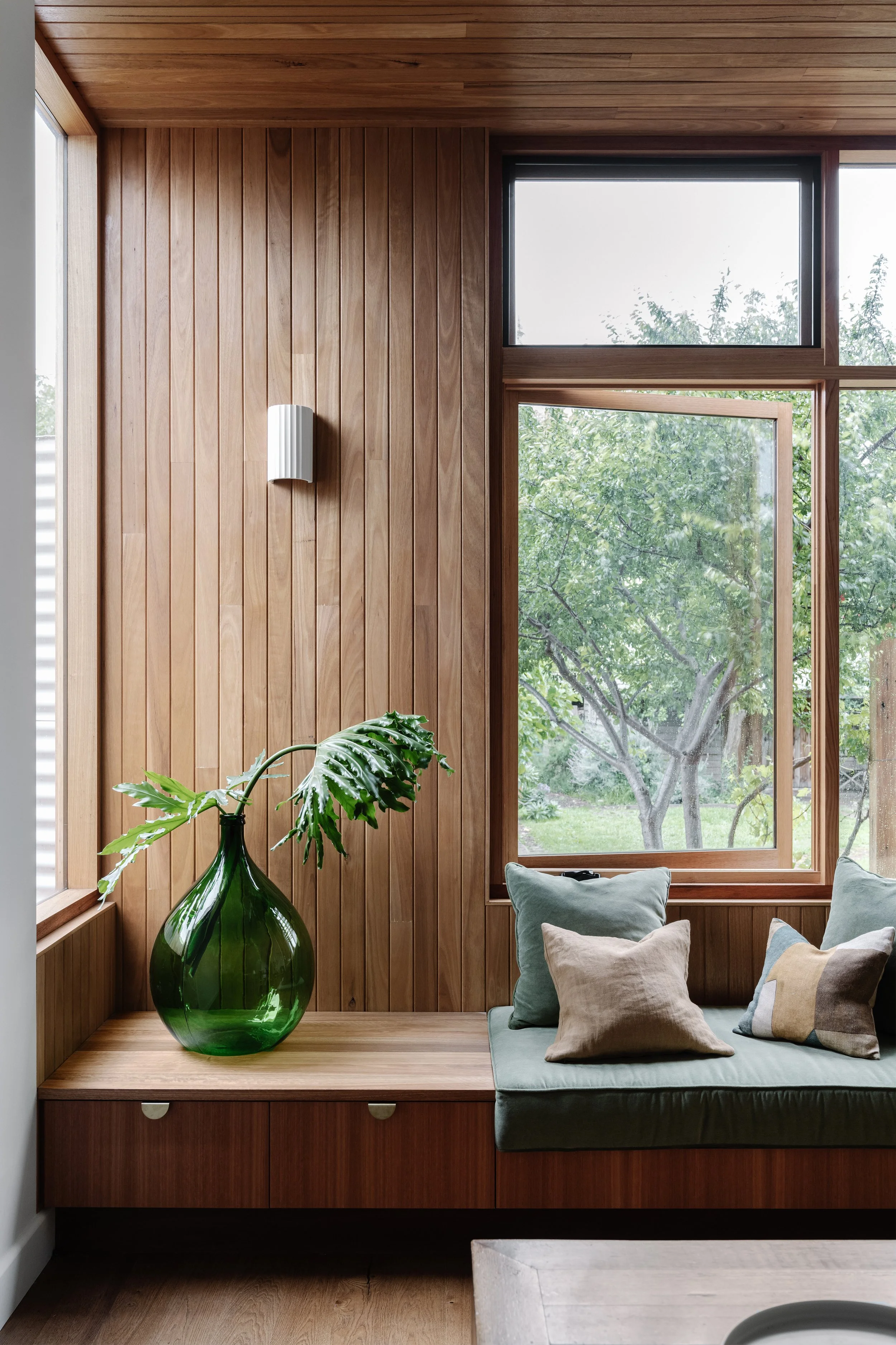 Interior of a room with wood-paneled walls and large windows showing green trees outside, a green glass vase with a leafy plant, and a cushioned bench with pillows.