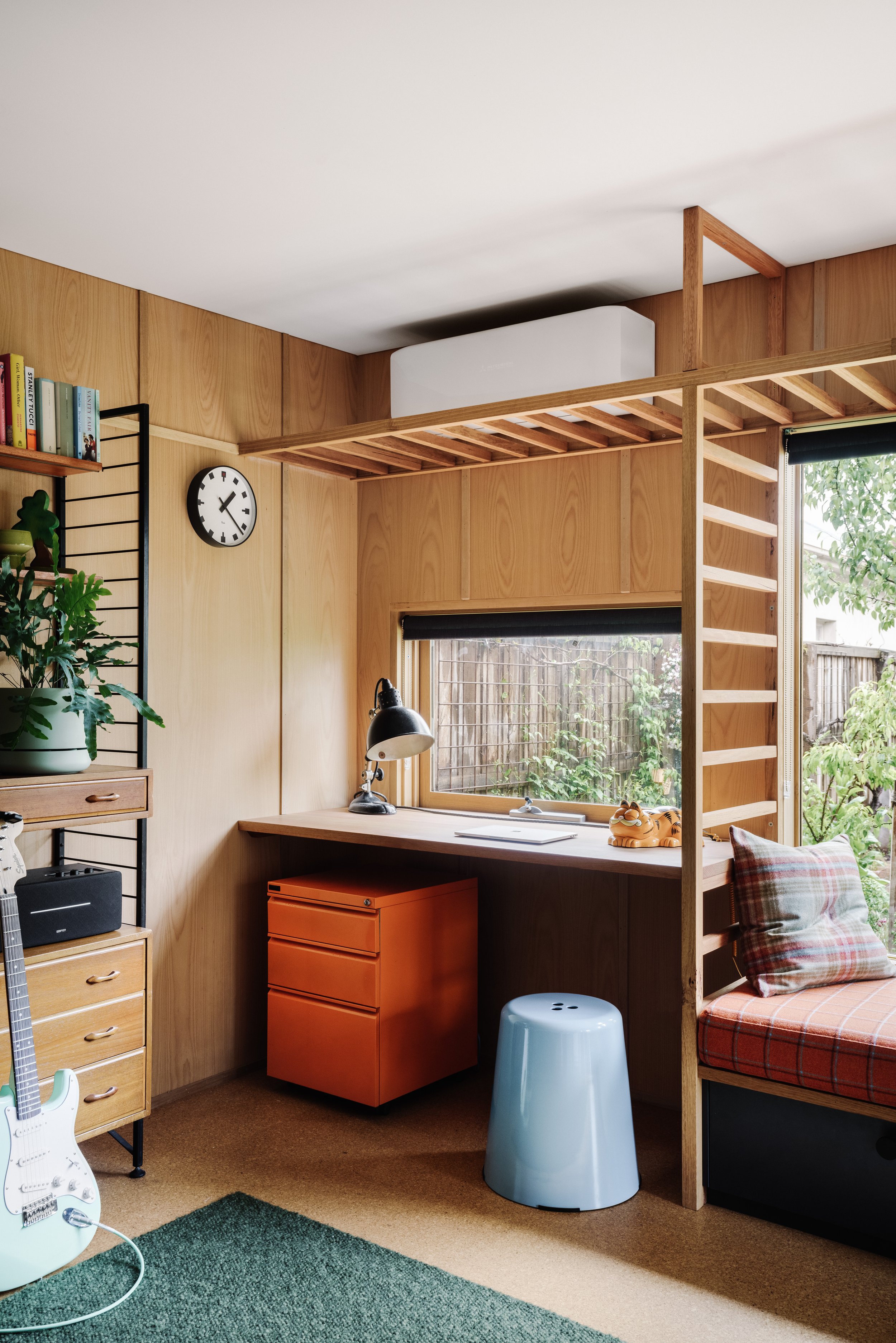 A cozy room with wooden panel walls, a small built-in desk space near a window, a white clock on the wall, a black desk lamp, and a bed with a plaid pillow. Decor includes a potted plant, books, and an electric guitar on a stand. There is a blue stool and an orange cabinet under the desk.