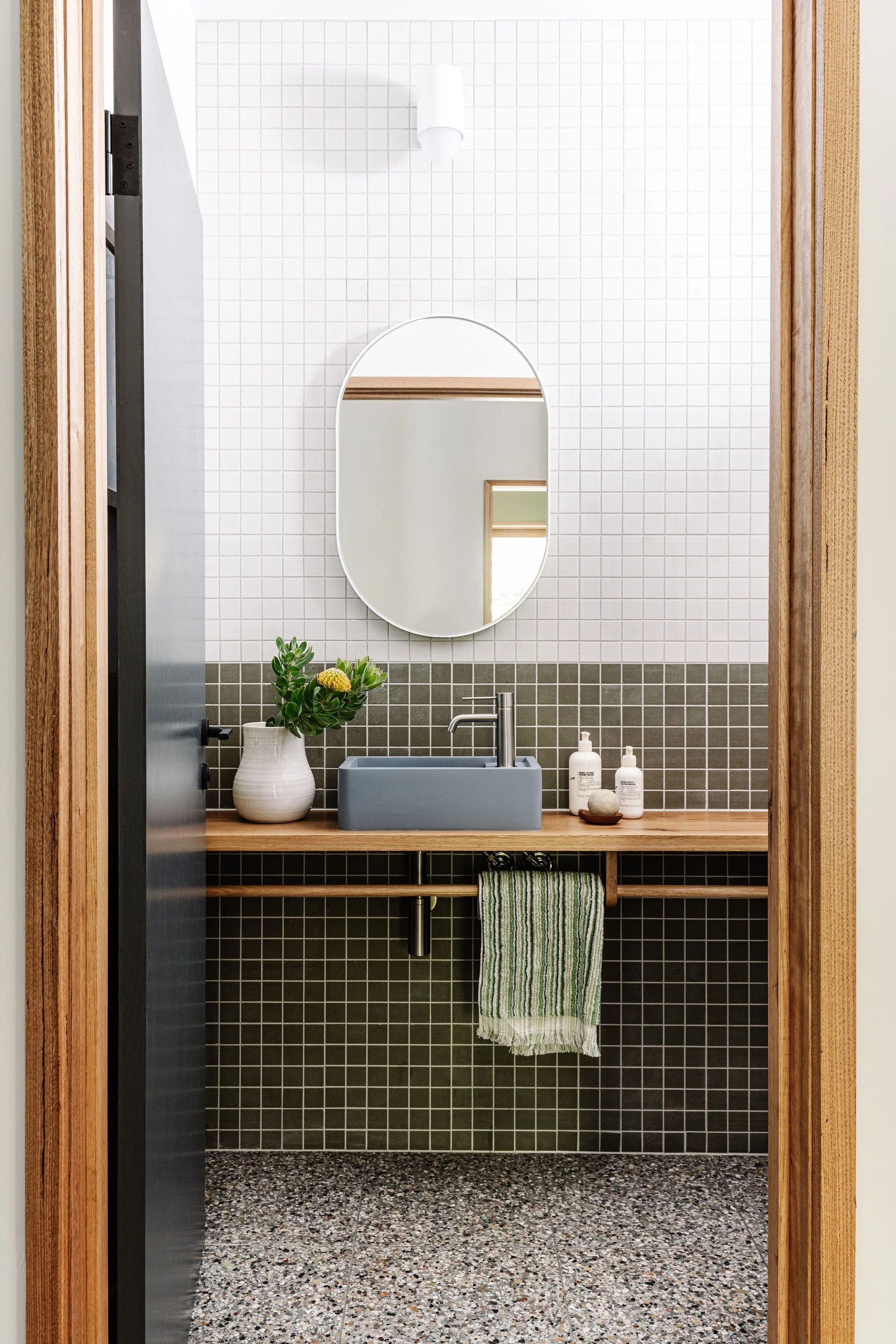 Modern bathroom with a small sink, a mirror, green and white striped towel, potted plant, soap bottles, and a wood shelf, with tile walls and a speckled tile floor.