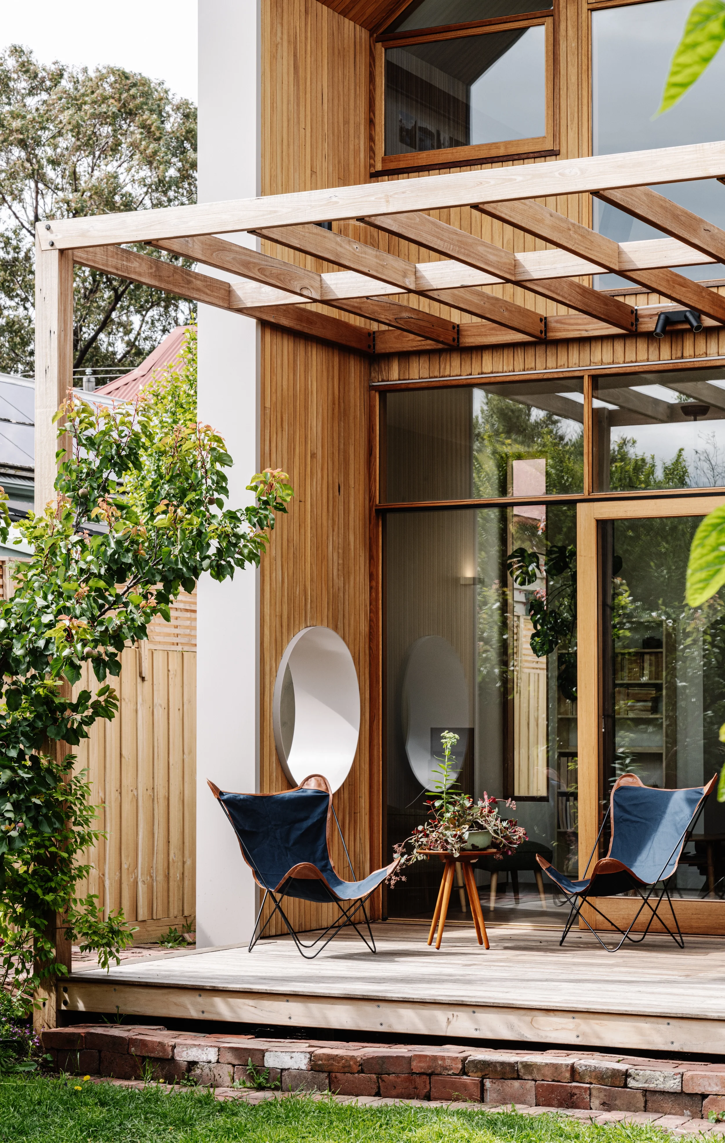 A modern wooden deck with two blue chairs and a small round table, decorated with plants, in front of a house with large glass windows and wooden exterior panels, surrounded by greenery.