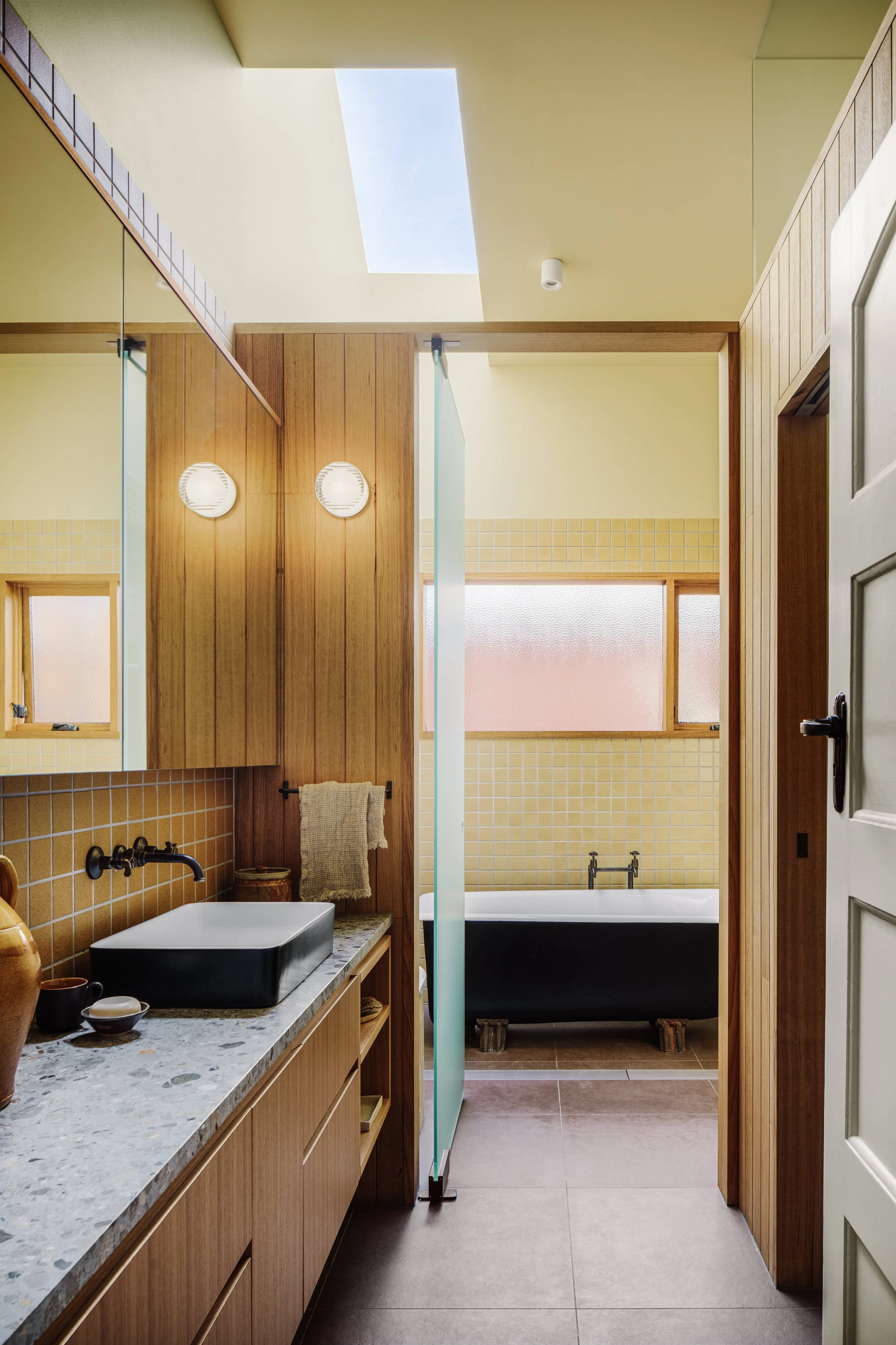 Bathroom with wooden cabinets, a black sink, and a bathtub at the back, separated by a glass divider, with a skylight and windows letting in natural light.