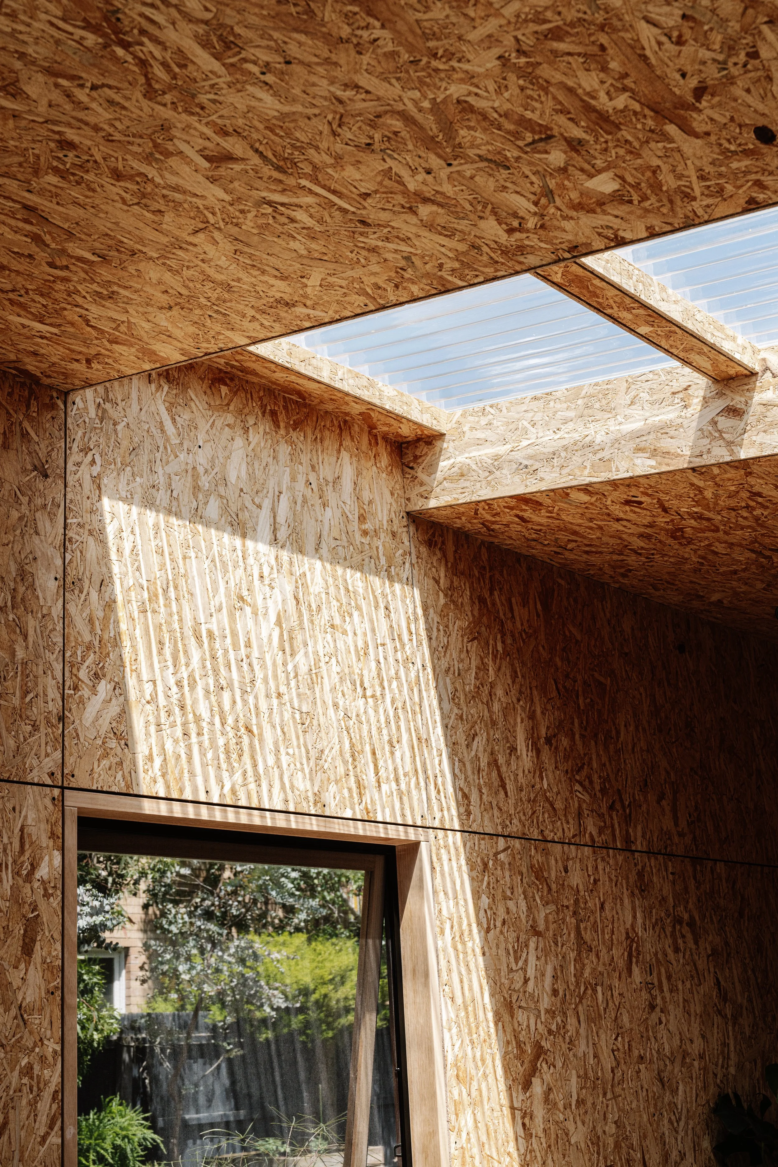 Interior view of a room with walls and ceiling covered in oriented strand board (OSB) panels, and a window showing a garden outside.