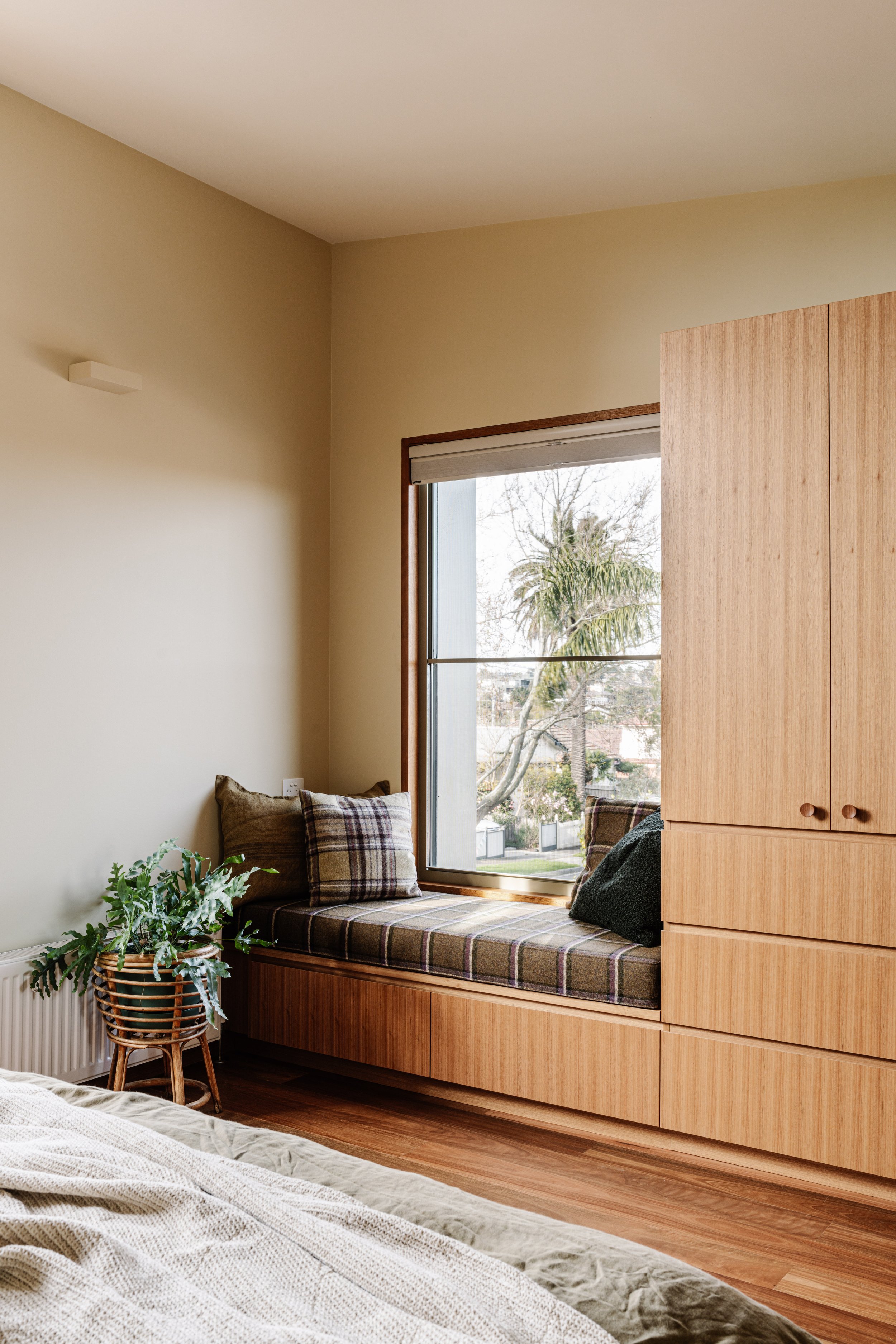A cozy nook by a large window with a built-in wooden bench and plaid cushions. A potted plant is on a stand next to the bench. Outside, a tree and palm are visible. The room has beige walls, wooden flooring, and a wardrobe partially shown on the right.