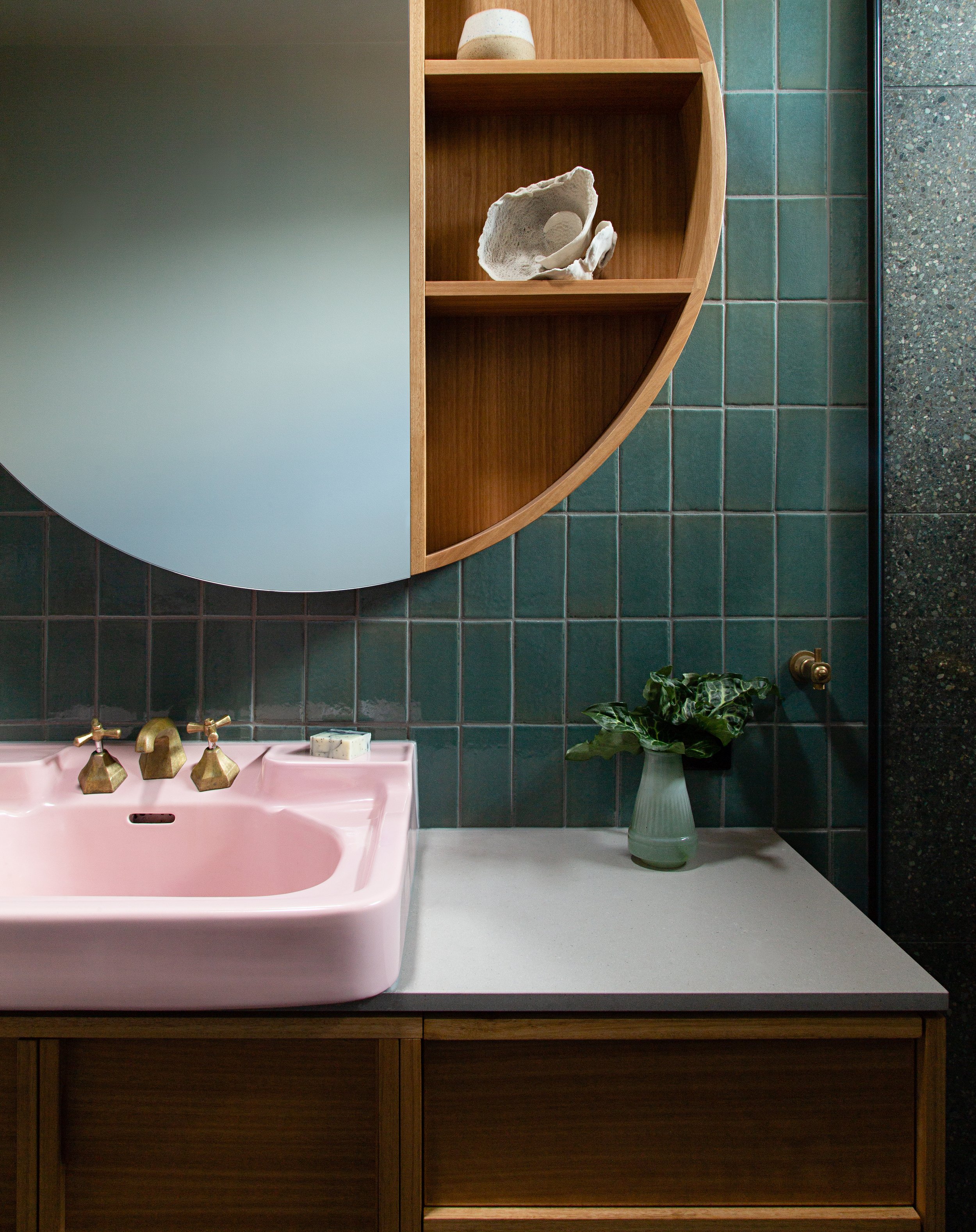 A pink vintage-style bathroom sink with brass fixtures, a rectangular marble soap dish, and a green plant in a clear vase on the white countertop. The background features dark green tiled walls and a wooden countertop with drawers.
