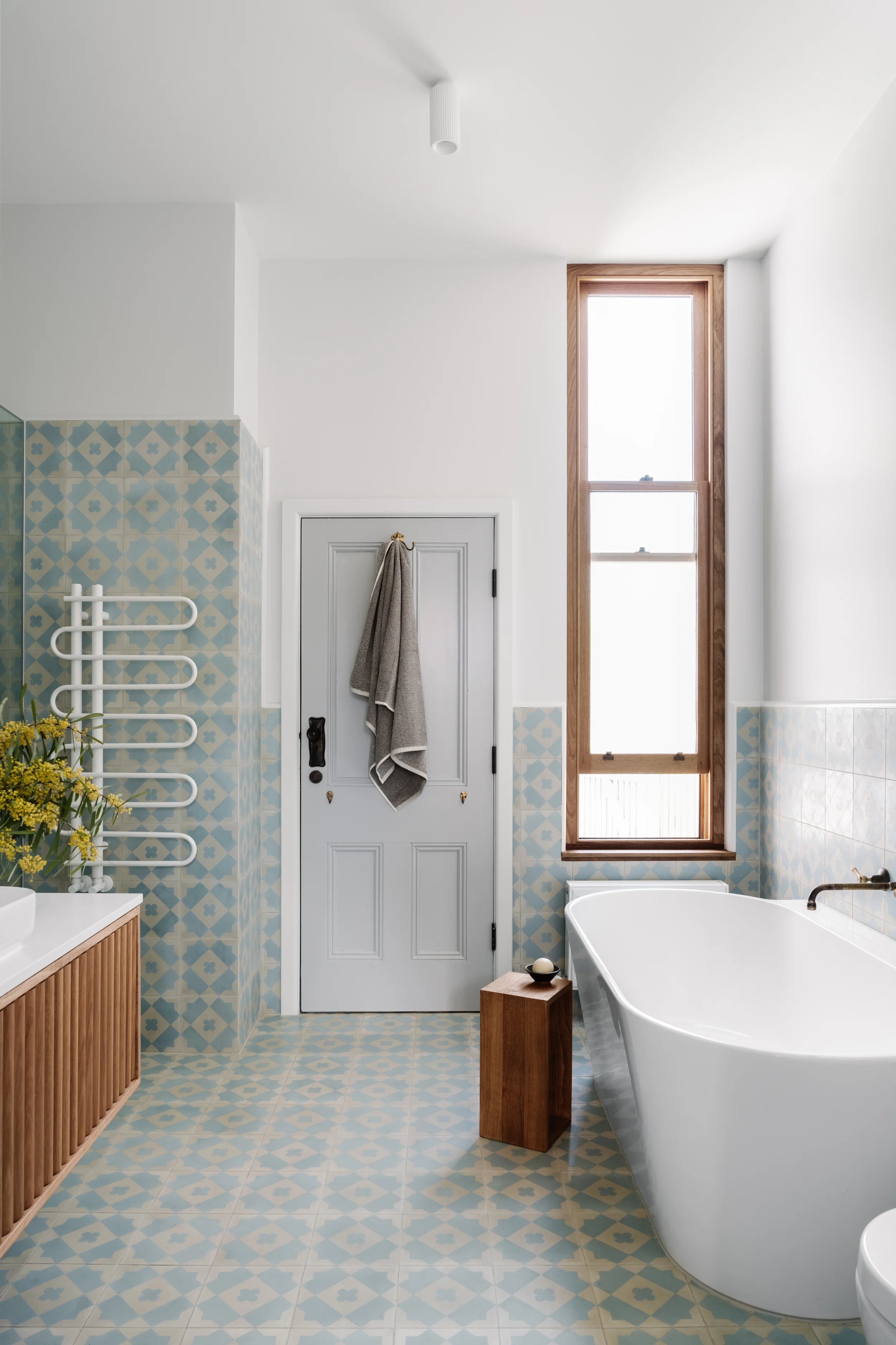 Modern bathroom with a freestanding bathtub, a tall narrow window with a wooden frame, a door with a towel hanging on it, blue patterned tiles on the walls and floor, and a wooden cabinet with a flower in a white bowl.
