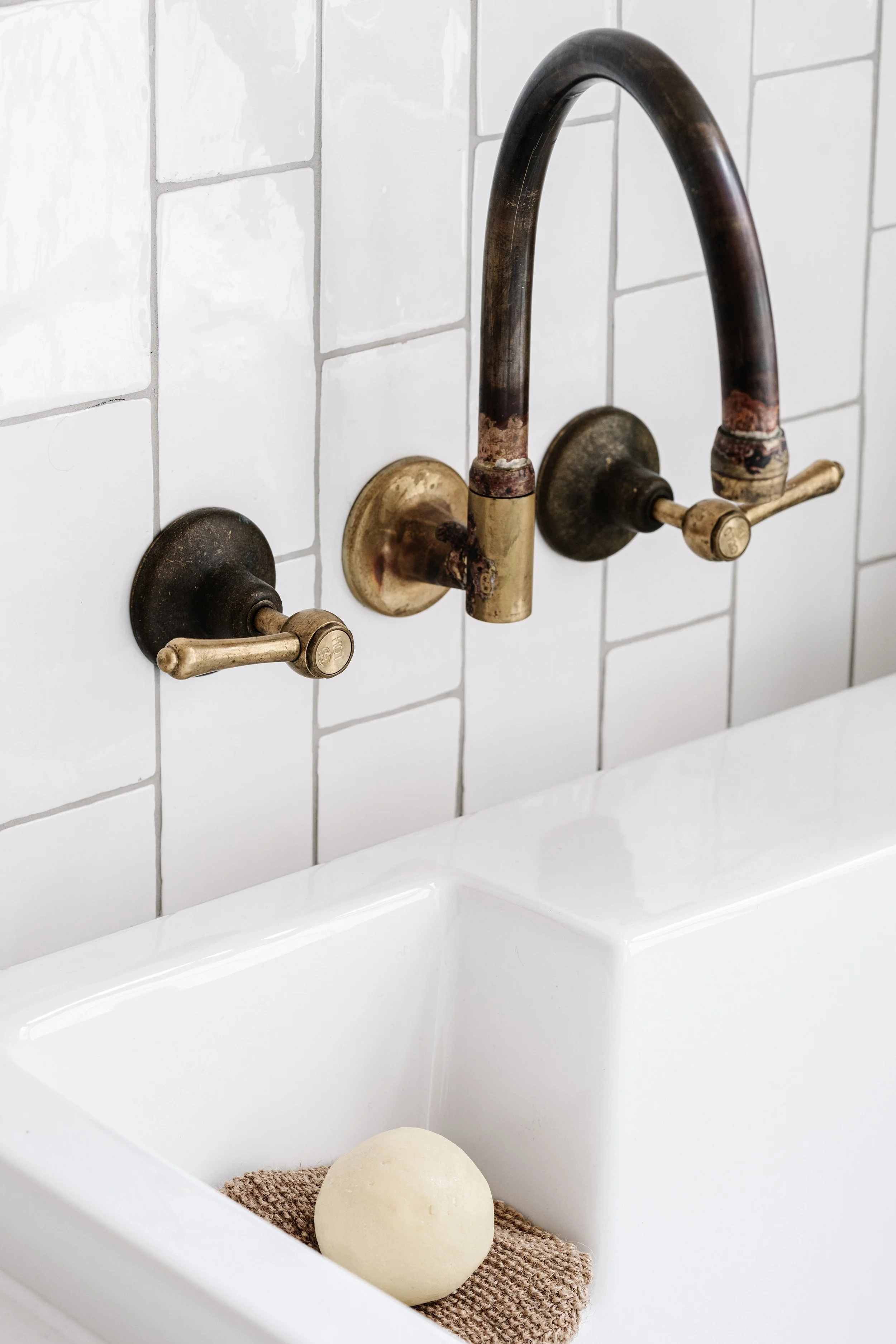 Close-up of an aged, rusty brass kitchen faucet mounted on a white tiled wall above a white sink. A round bar of soap rests on a small burlap towel inside the sink.
