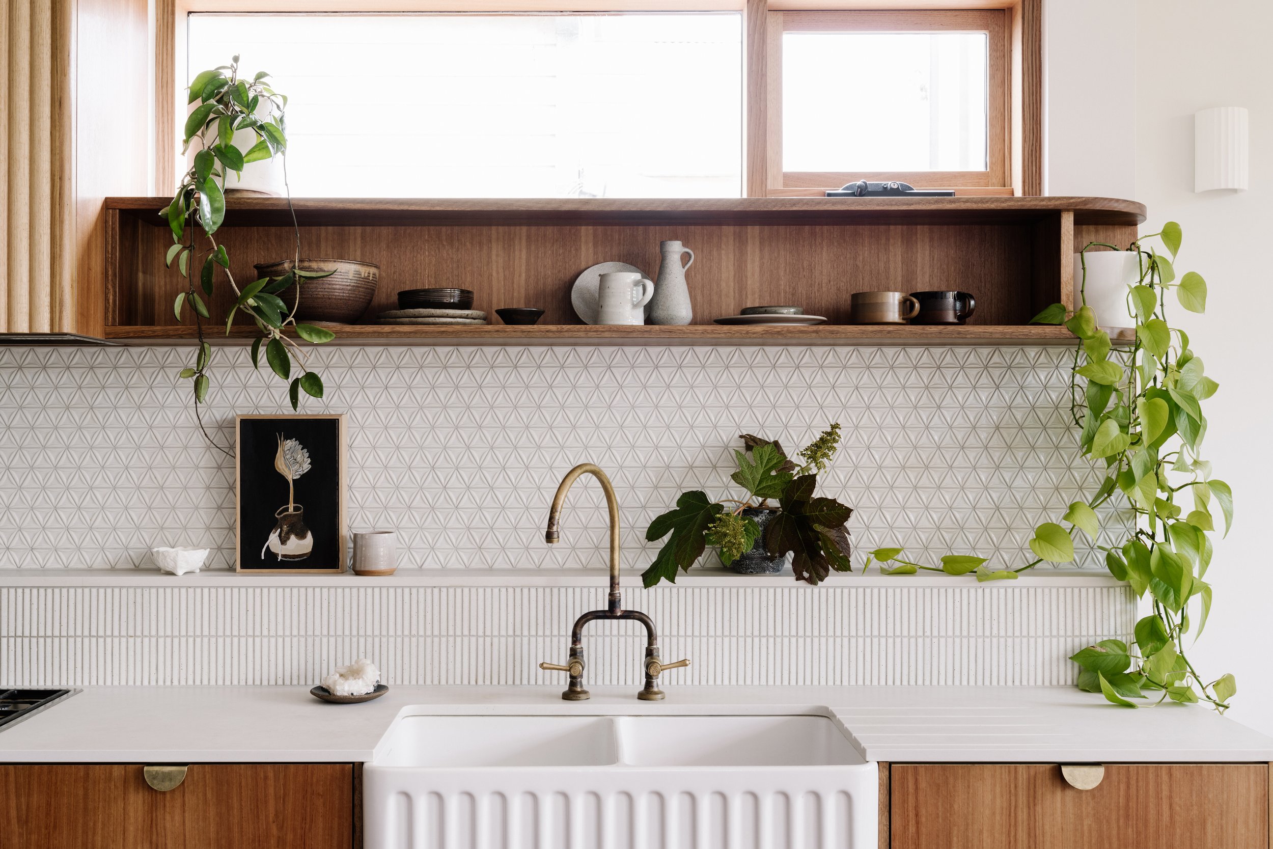 A kitchen with a white sink, wooden cabinetry, and a patterned backsplash. Decorative plates and ceramics are on a wooden shelf above the sink, with potted plants on either side. A window above the shelf brings in natural light.