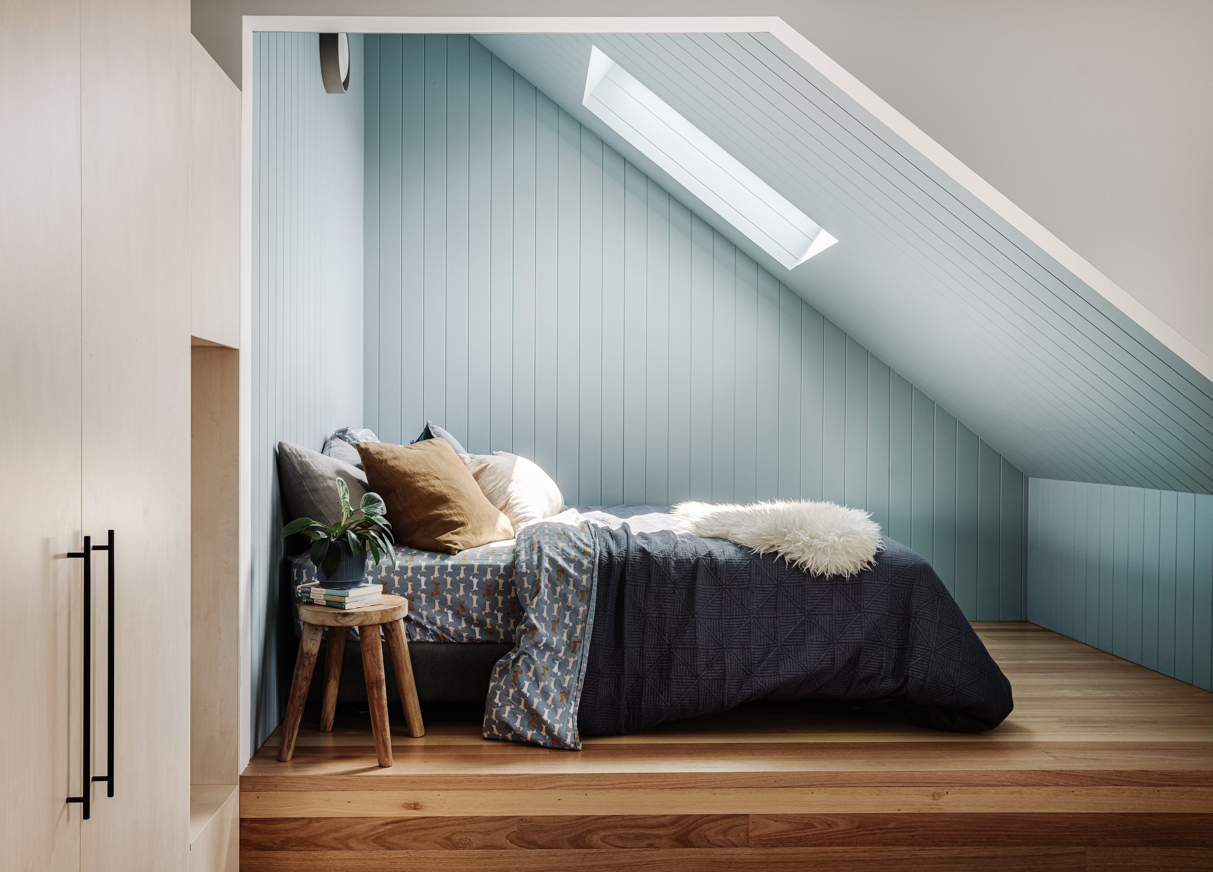 A cozy bedroom with a sloped ceiling and a skylight window, featuring a bed with pillows and a blanket, a small wooden stool with a stack of books and a potted plant, and a pale blue paneled wall.