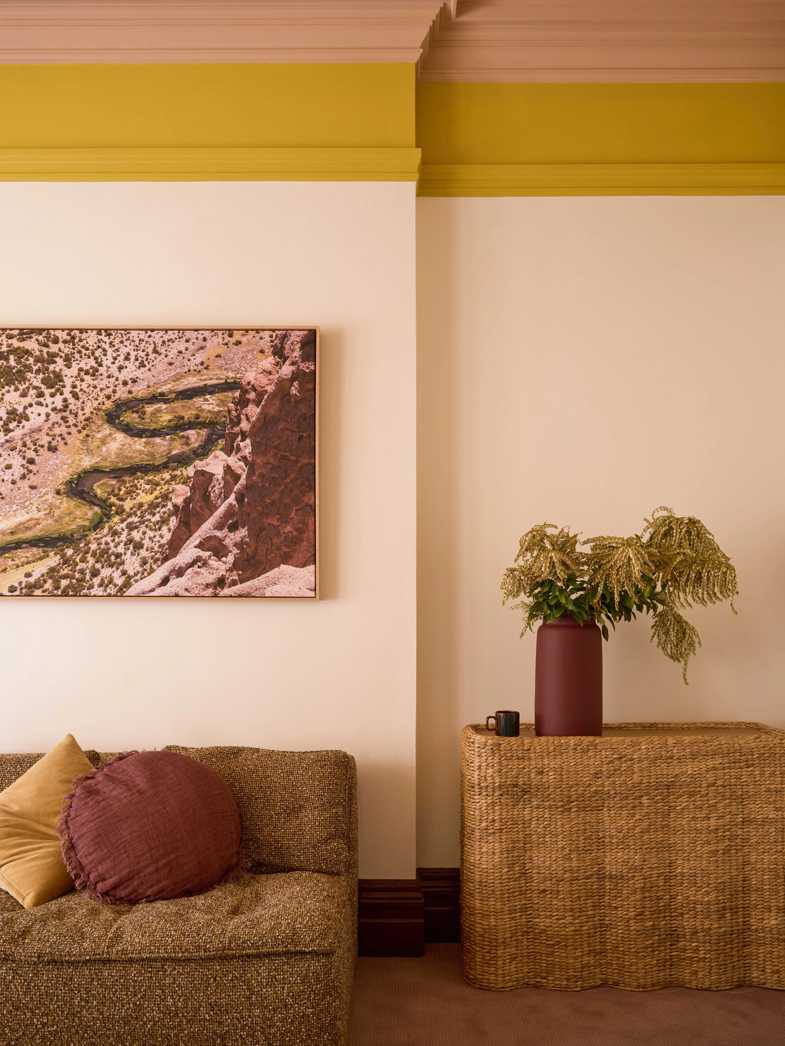 Living room corner with a sofa, pillows, a framed landscape photograph on the wall, a large vase with flowers on a wicker cabinet, and a small cup.