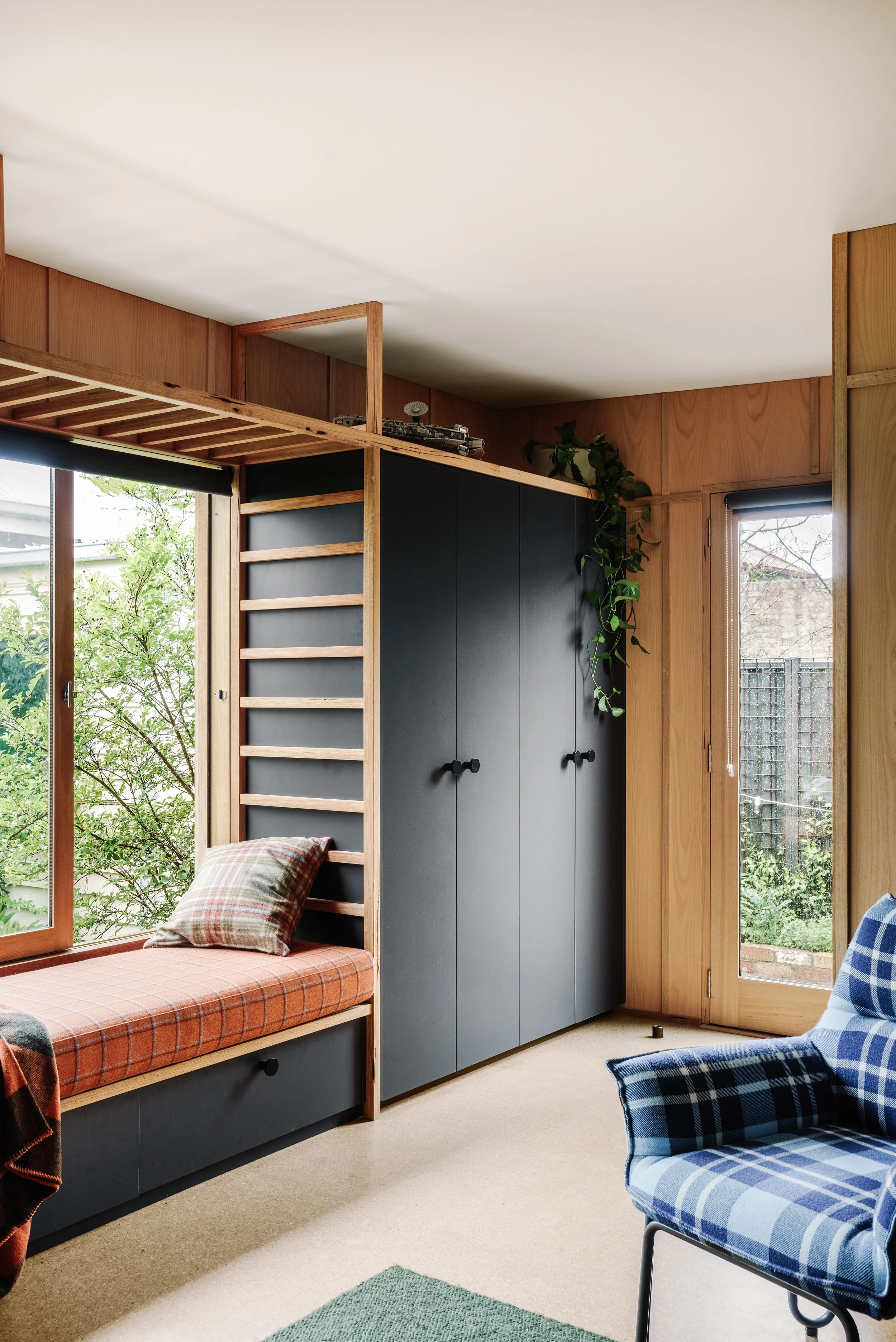 A cozy living room corner with a window seat featuring a plaid cushion and pillow, a tall black wardrobe with wooden accents, a green potted plant, and large windows showing a garden outside.