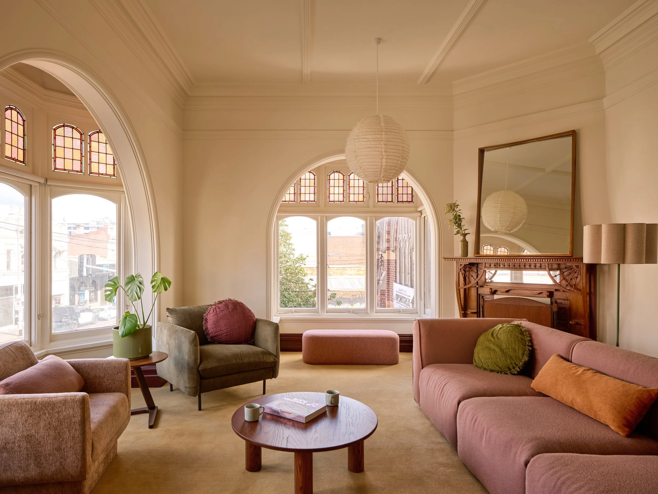 A cozy living room with several chairs and sofas in pastel pink, green, and beige, a round wooden coffee table with two cups and a magazine, large windows with stained glass, and a mirror on the wall reflecting a hanging paper lantern.