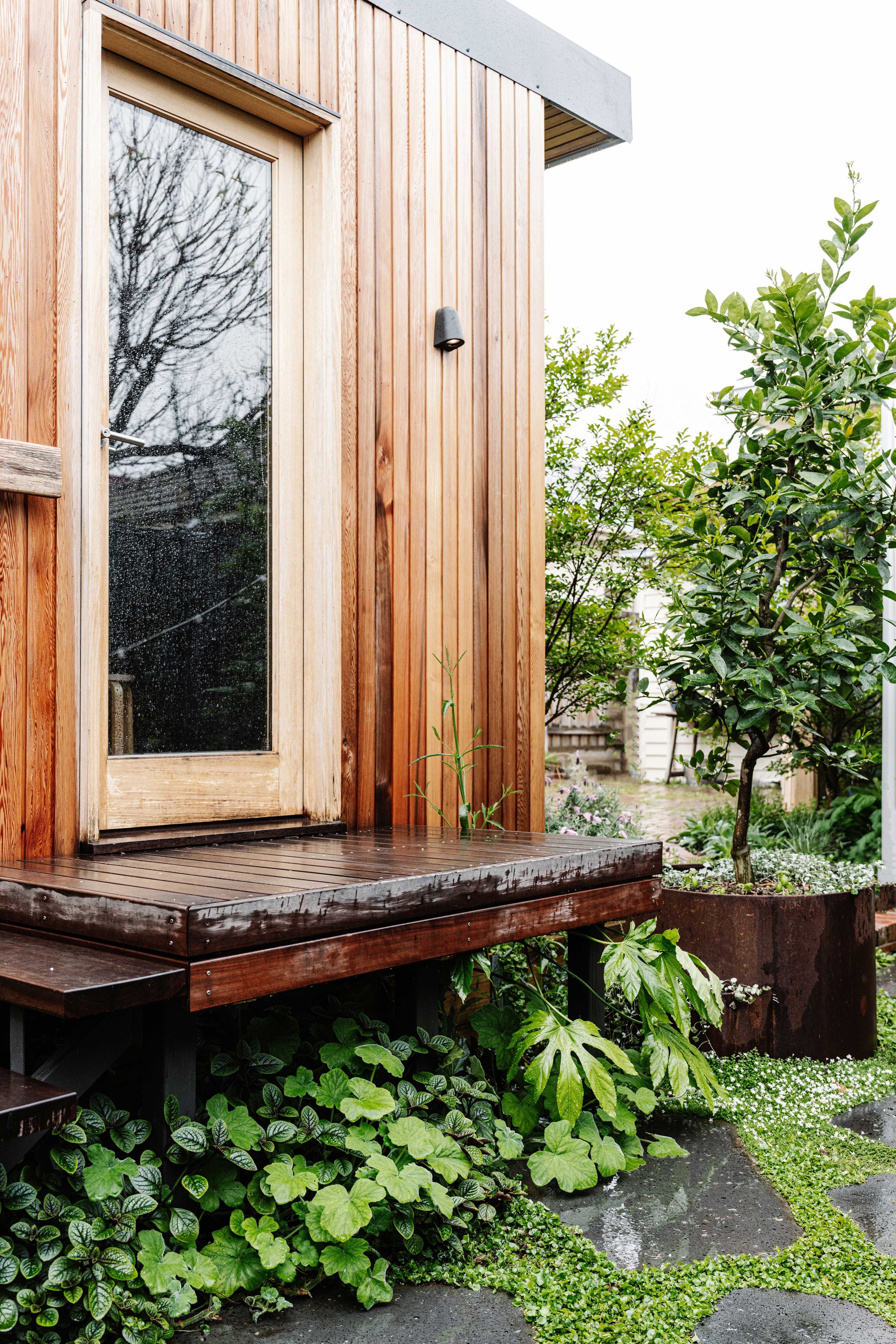 Wooden exterior door with glass panel on a deck, surrounded by lush green plants and shrubbery in a garden.