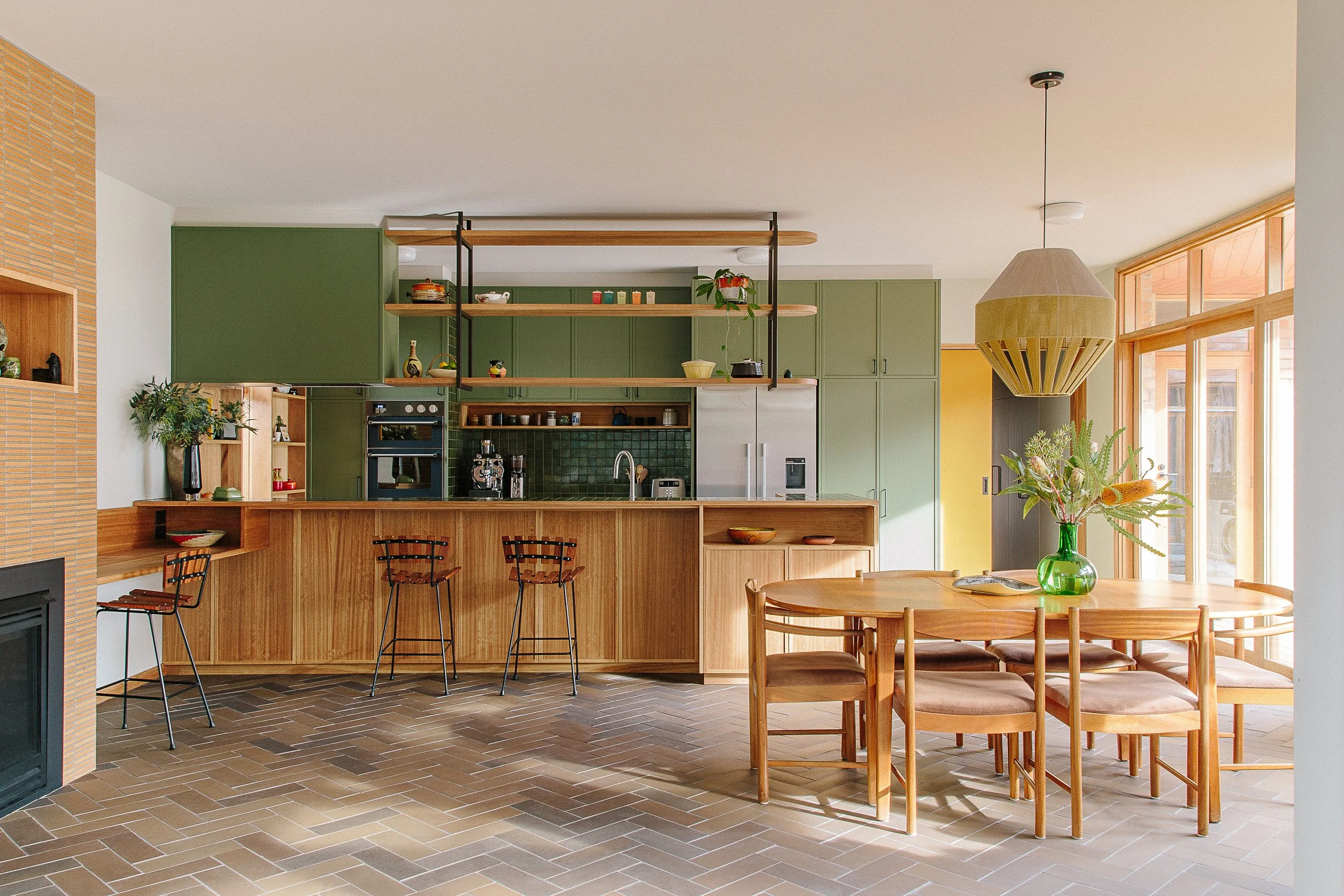 Open-plan kitchen and dining area with green cabinets, wooden accents, a hanging light fixture, and a large window with blinds