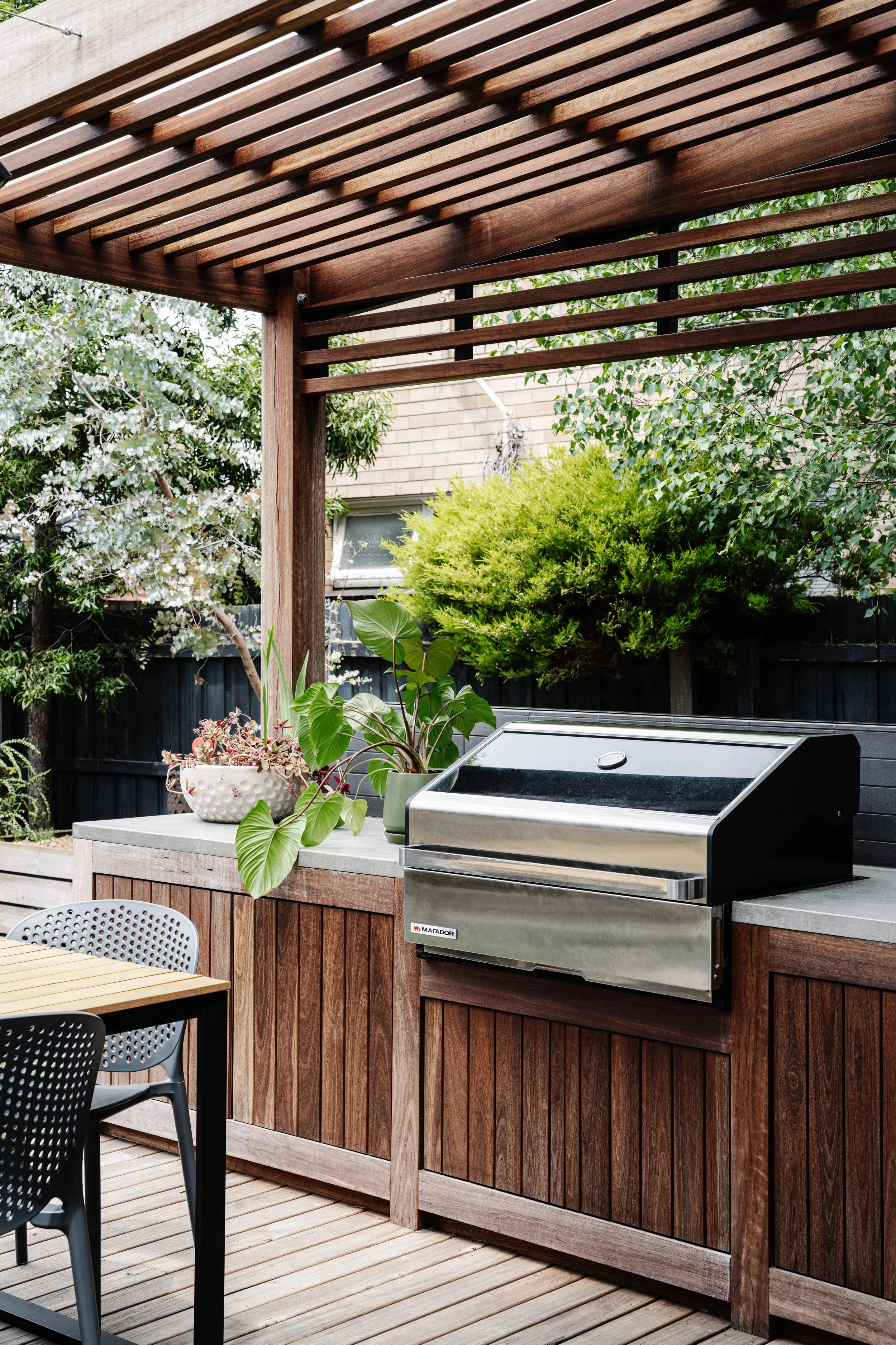An outdoor kitchen area with a stainless steel grill, potted plants, and a wooden countertop beneath a slatted wooden pergola, surrounded by trees and a brick wall.