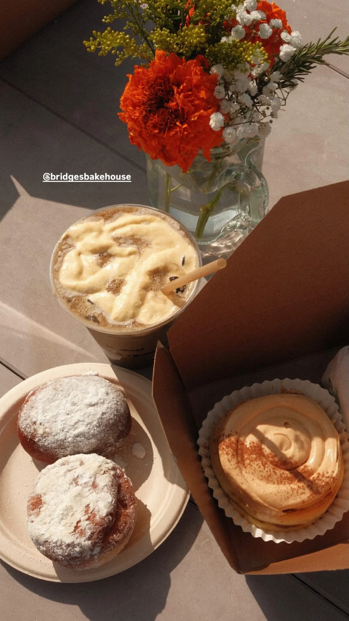A table with a vase of orange, white, and yellow flowers, a cold drink topped with whipped cream, two powdered sugar dusted muffins on a paper plate, and a lemon-flavored cupcake with a dusting of paprika in a cardboard box.