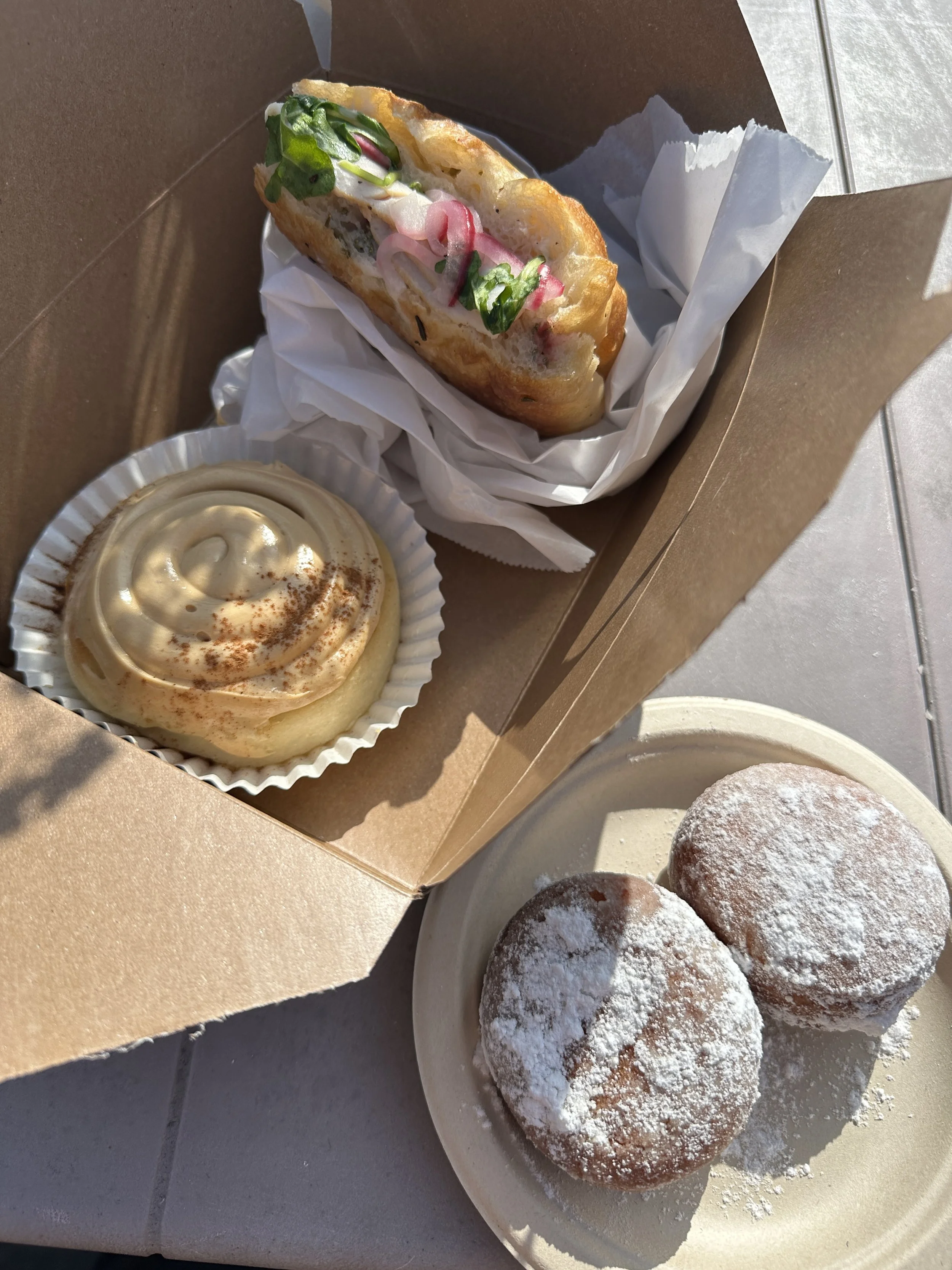 A food presentation with a hot dog topped with green onions and other garnishes inside a brown takeout box. A cup of vanilla soft-serve ice cream with cinnamon sprinkles. Two powdered sugar-dusted fried doughnuts on a white plate.