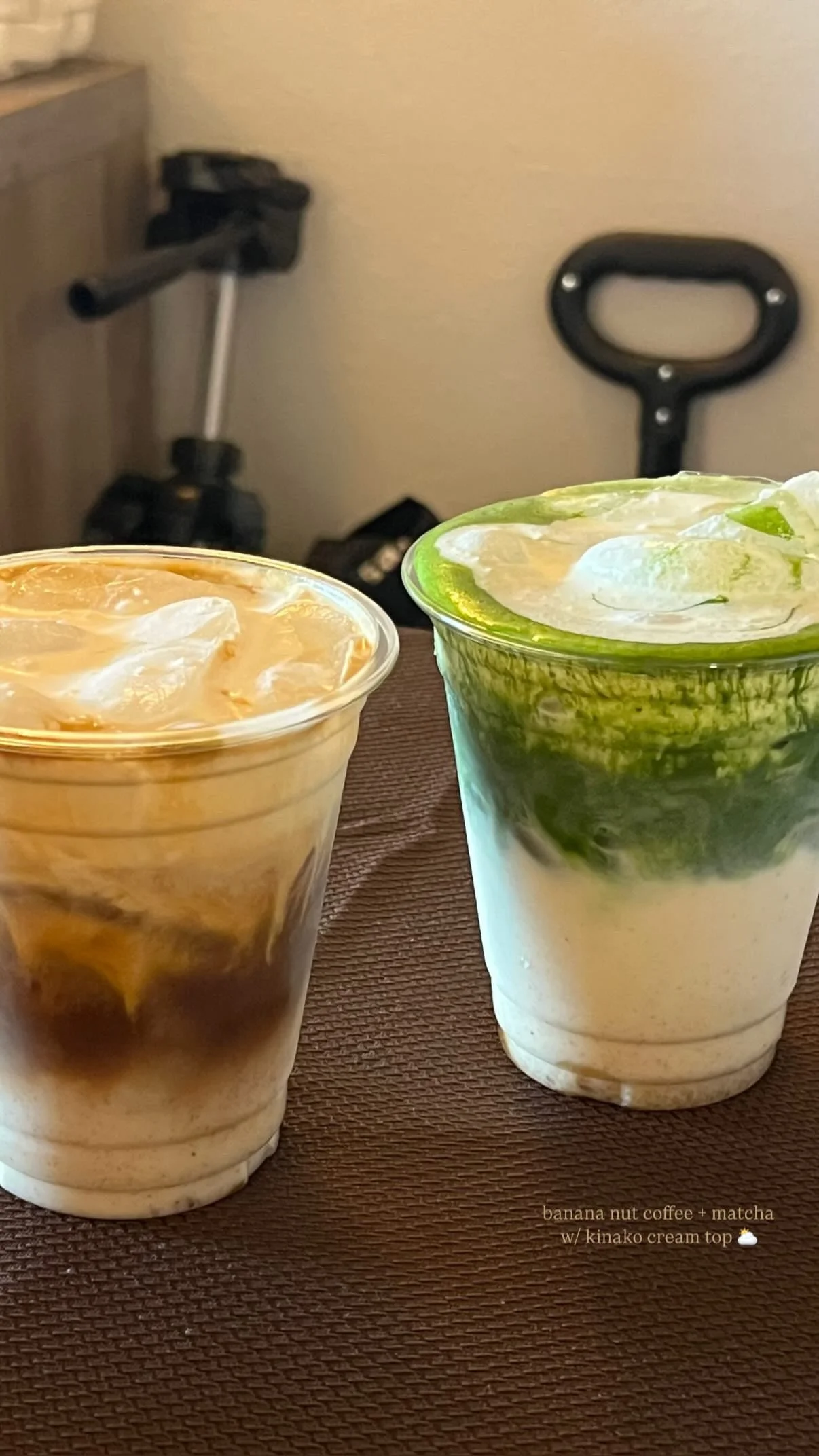Two cups of iced beverages on a brown surface, one with a caramel-colored drink and the other with a green matcha layer, with a background of a scooter and a black handle tool.