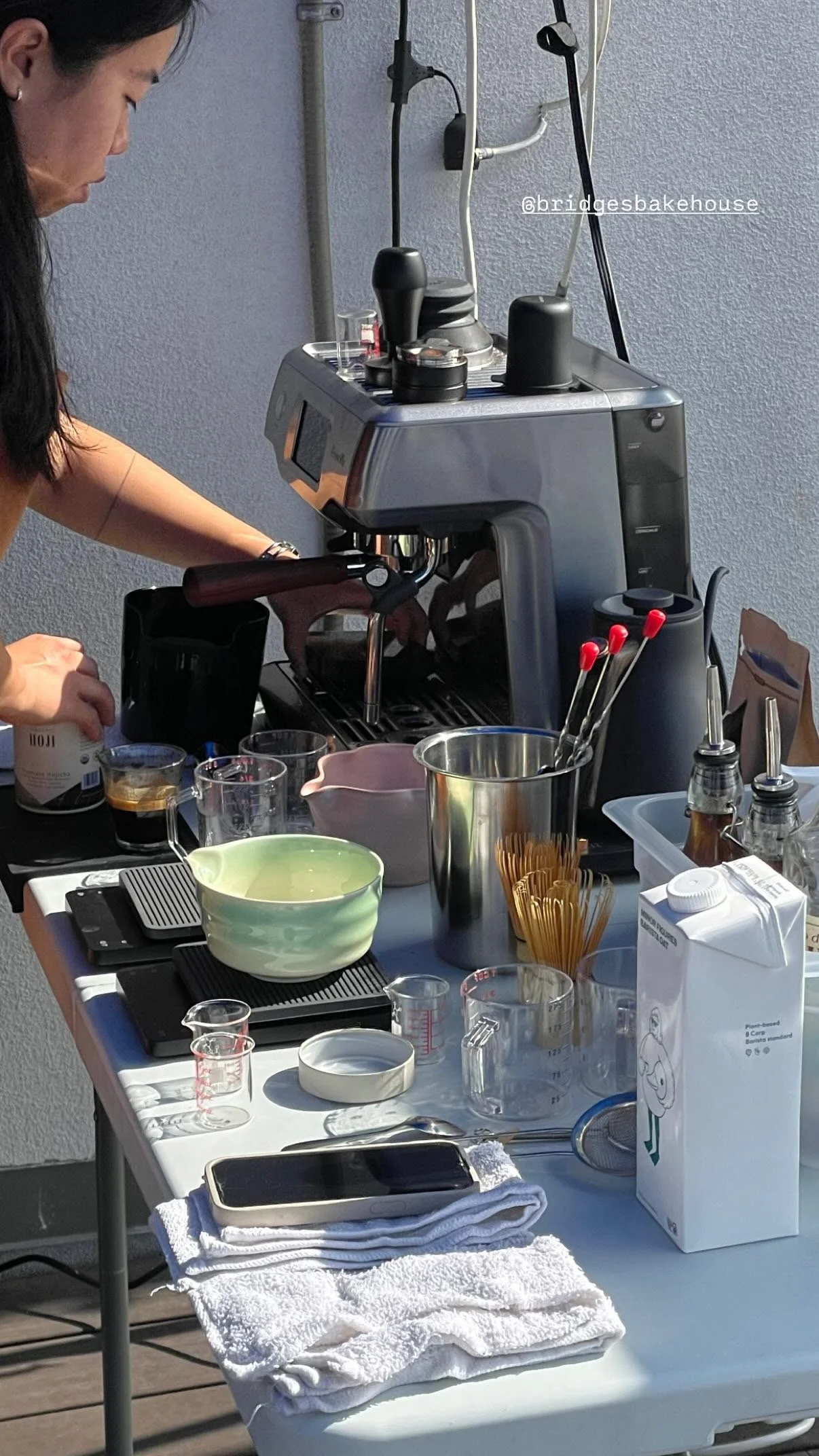 A coffee brewing setup on a table with various tools and ingredients, including an espresso machine, cups, measuring cups, utensils, a milk carton, and a towel, with a person working in the background.