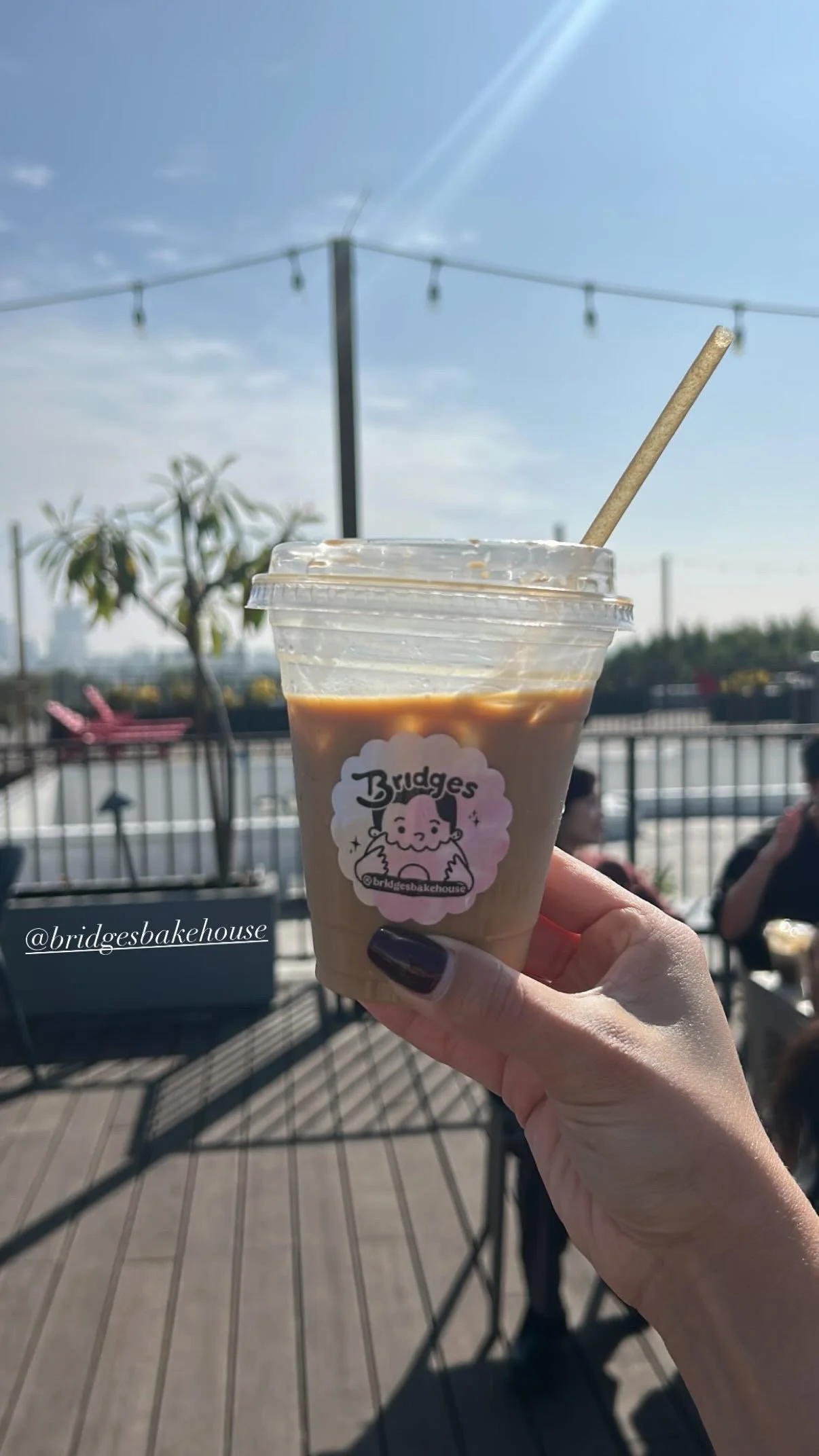 A hand holding a plastic cup with iced coffee on an outdoor patio with string lights overhead and blurred people in the background.