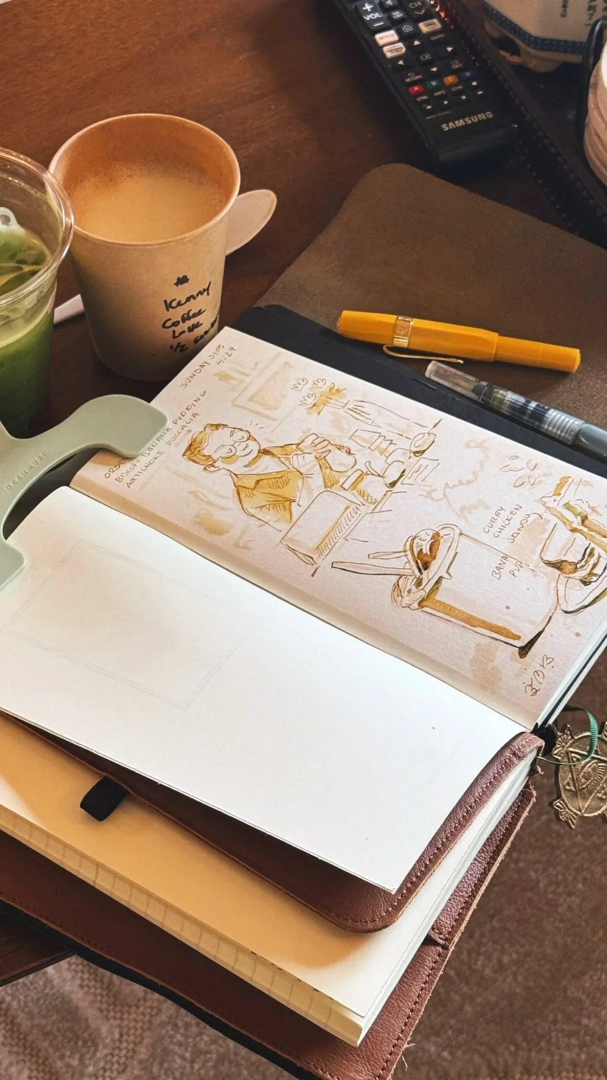 A sketchbook open to a page with a hand-drawn cartoon of a person sitting at a desk, holding a coffee cup, with a spoon and dessert in front. There are two coffee cups, pens, a remote control, and a stack of notebooks on a wooden table.