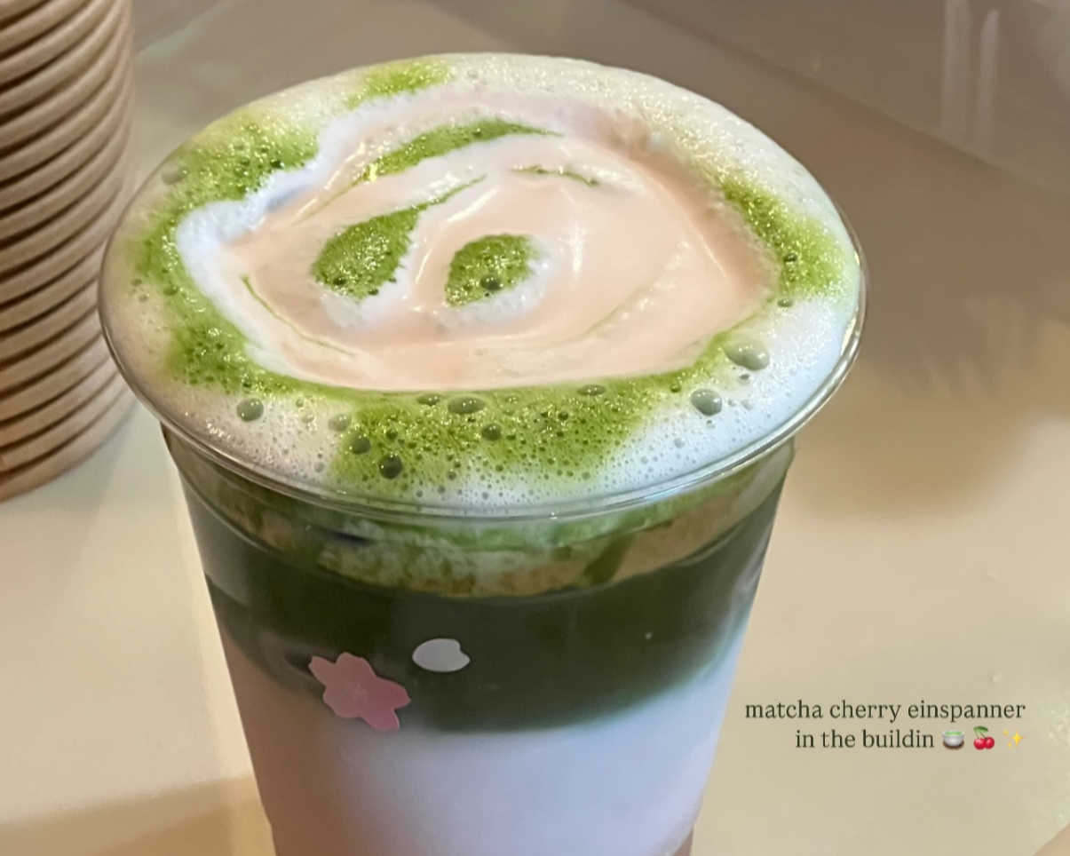 A matcha cherry drink in a tall glass with pink cherry blossom design, topped with whipped cream and green matcha swirl.