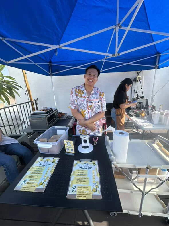 A smiling man standing behind a table with yellow and white menus, under a blue canopy, with a woman working at a coffee station in the background.