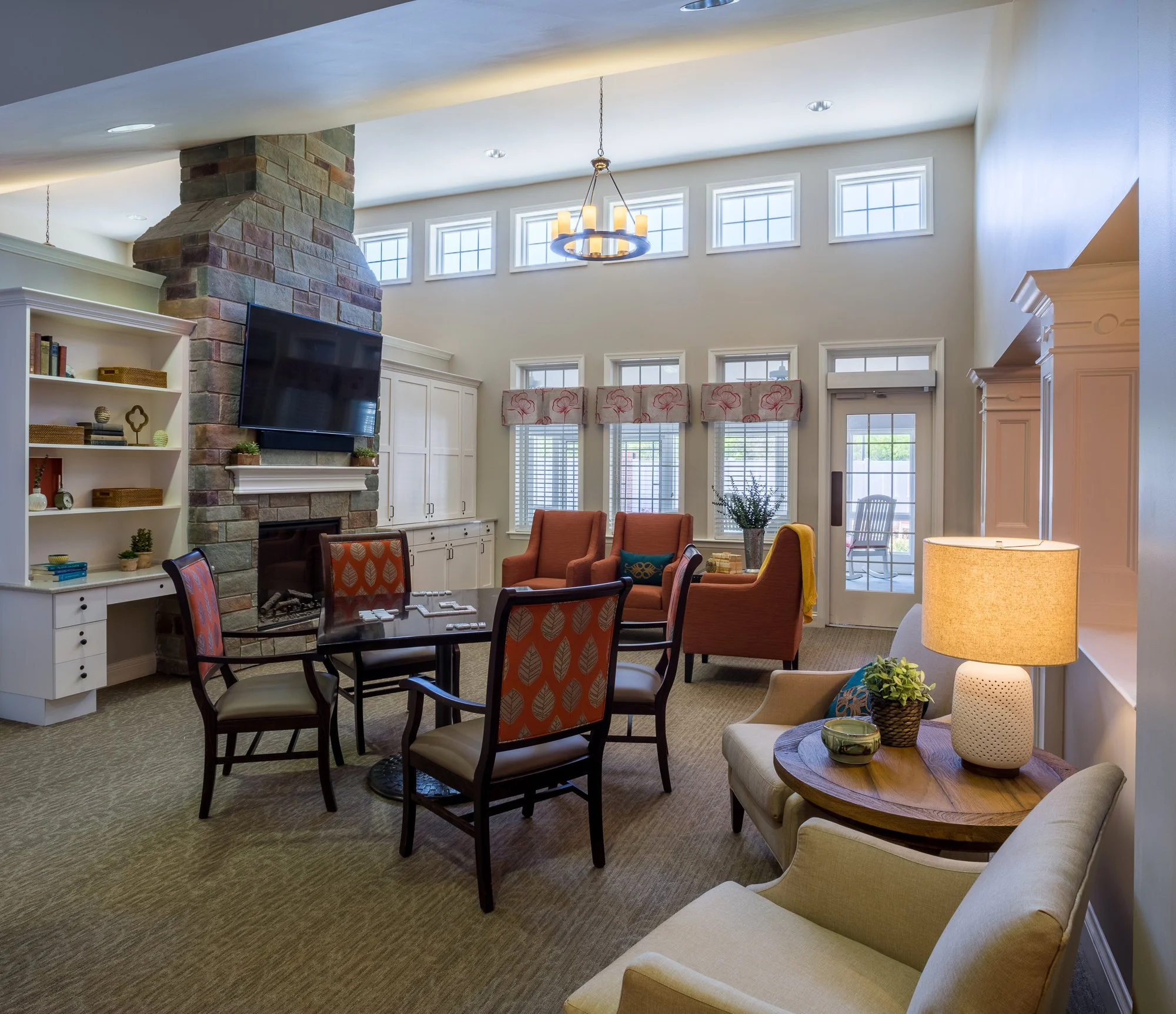 A living room with a stone fireplace, a mounted flat-screen TV, a white bookshelf, several chairs, a small round table with plants and decorative objects, and a wall with multiple small windows near the ceiling allowing natural light.