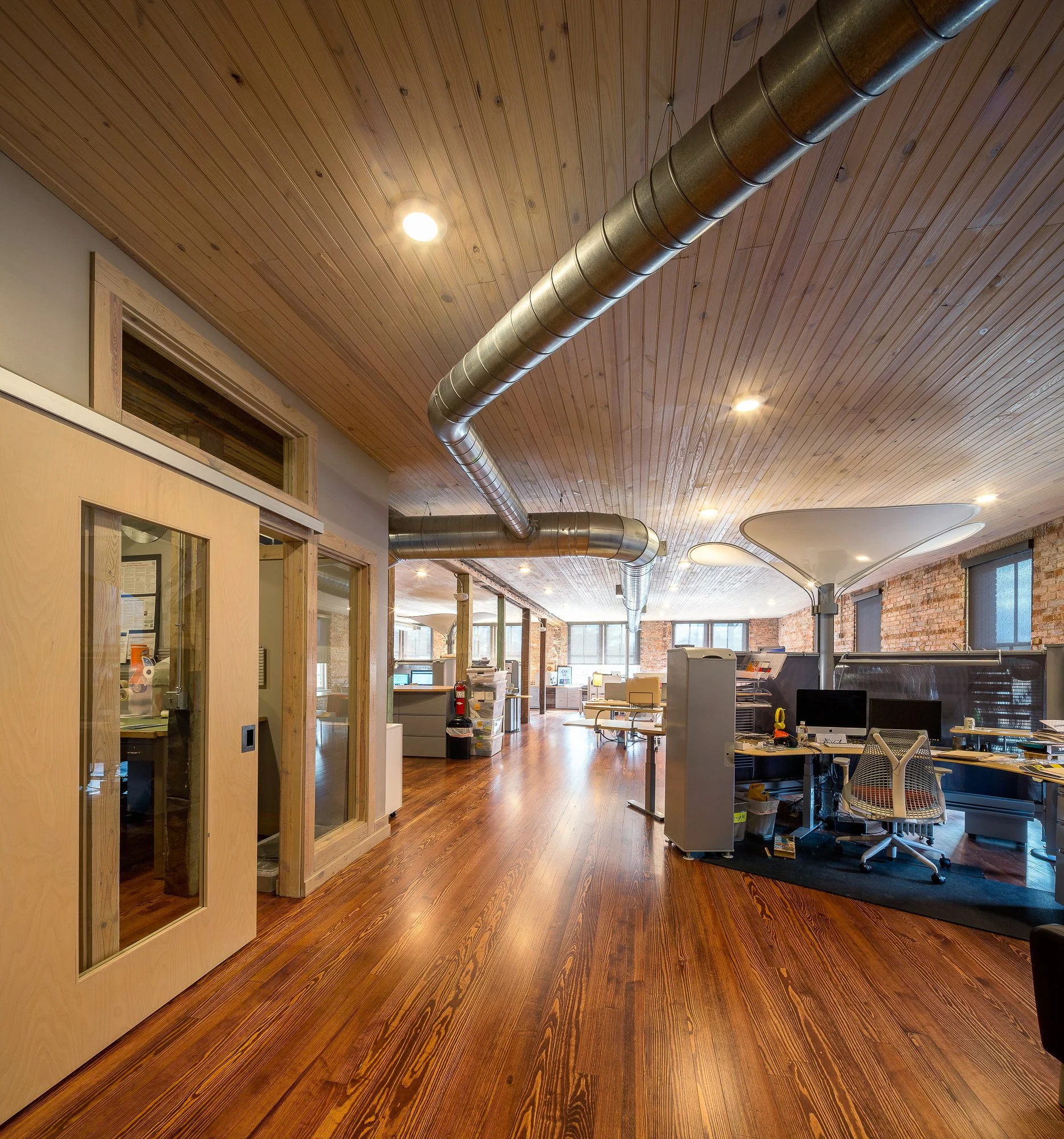 Interior view of a modern office space with wooden floors, wooden ceiling, exposed ductwork, and several workstations and cubicles.