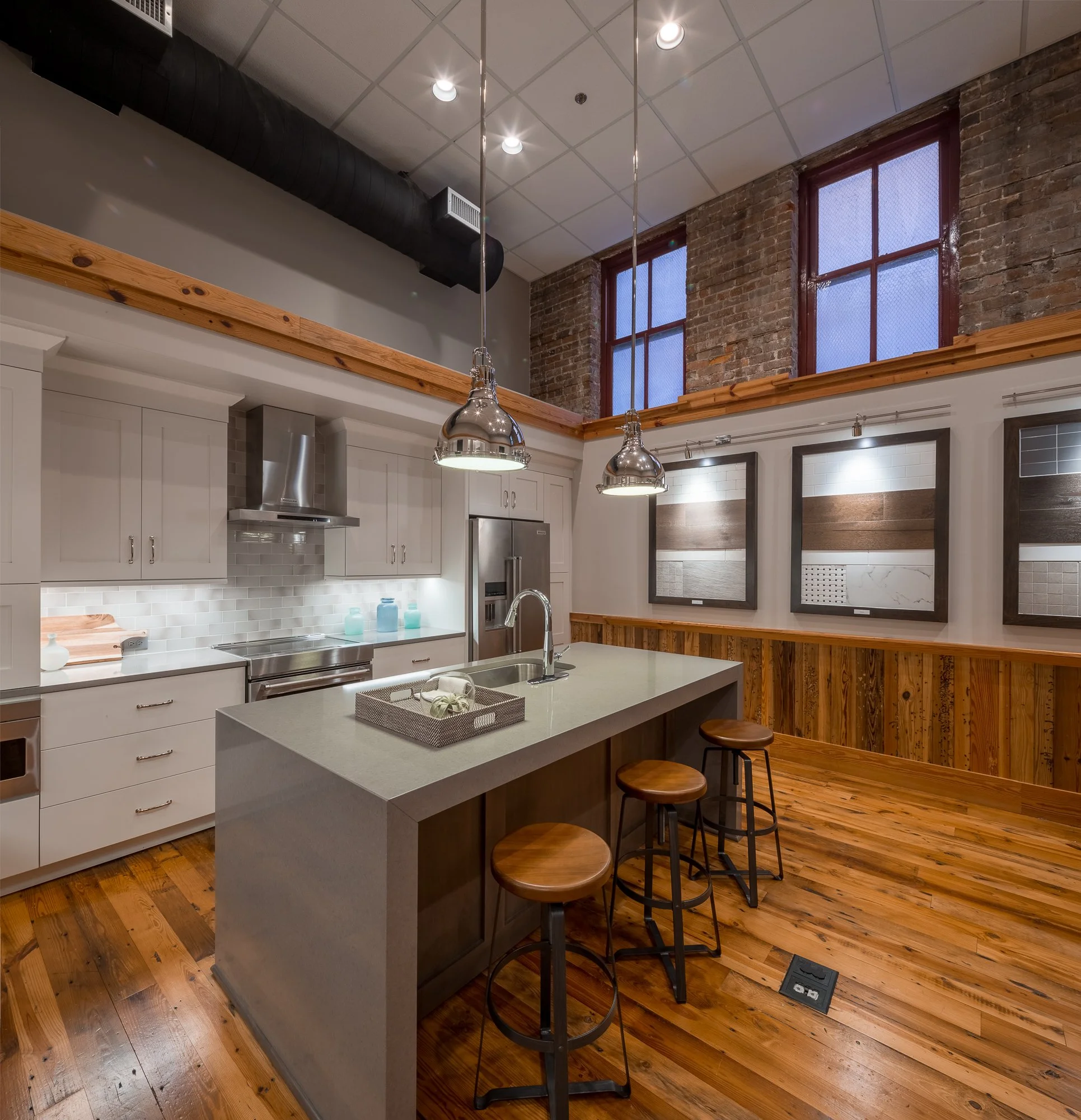 Modern kitchen with white cabinets, gray kitchen island, brick walls, exposed wooden beams, large windows, and wood flooring. Pendant lights hang over the island.