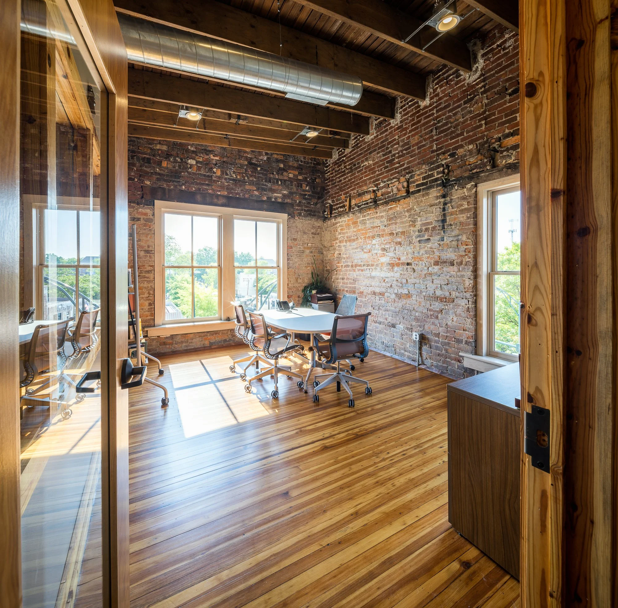 A room with exposed brick walls, wooden ceiling beams, and wood flooring. Large windows let in natural light. There is a conference table with chairs, a plant, and a cabinet near the window.