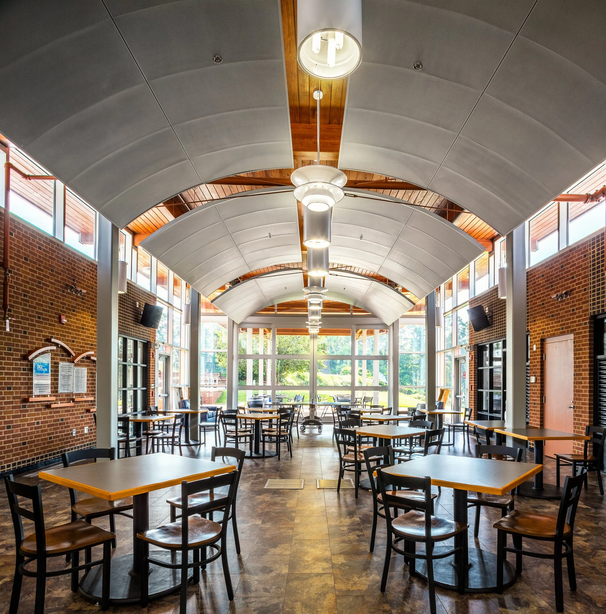Interior of a spacious building with a high, arched ceiling featuring large, modern pendant lights and wooden accents. The space has large windows along the walls allowing natural light, with brick walls and tables and chairs arranged for seating.