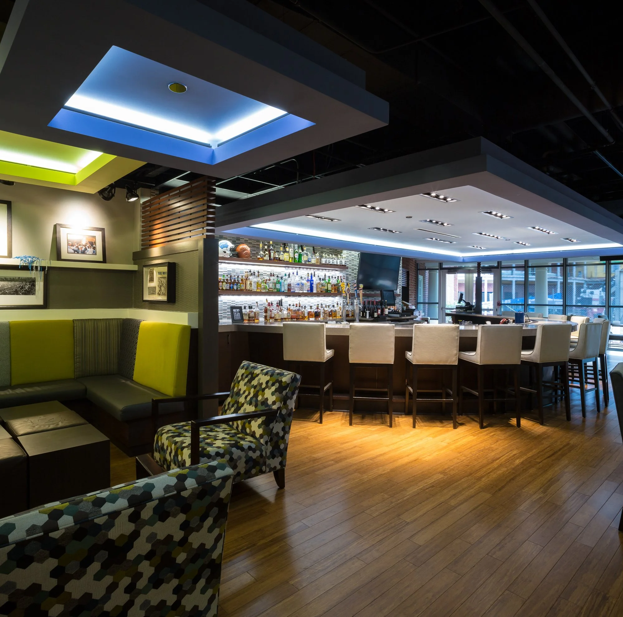 Interior view of a modern bar area with a ceiling featuring colorful, geometric-shaped light installations in blue, green, and white, with a variety of bottles on display behind the bar, and seating with patterned and solid-colored chairs.
