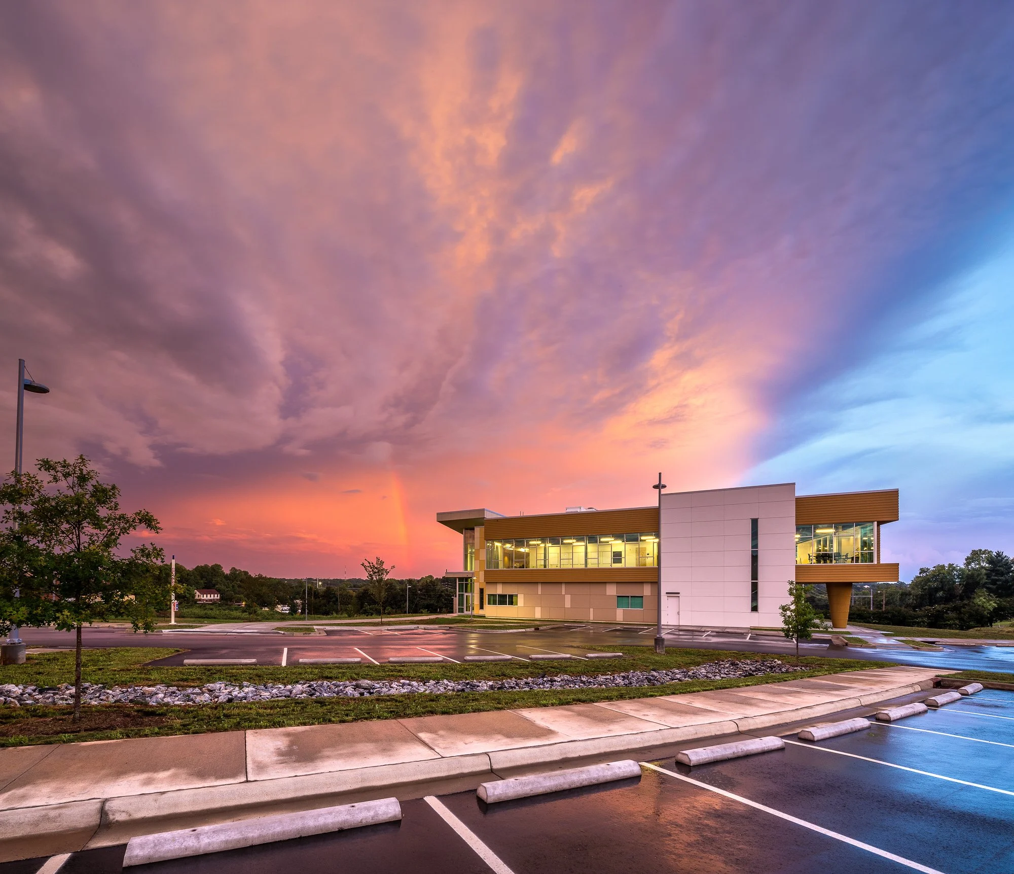 A modern, glass-fronted building in a parking lot during sunset, with pink and purple clouds in the sky, some trees, and an empty parking space with white parking blocks in the foreground.