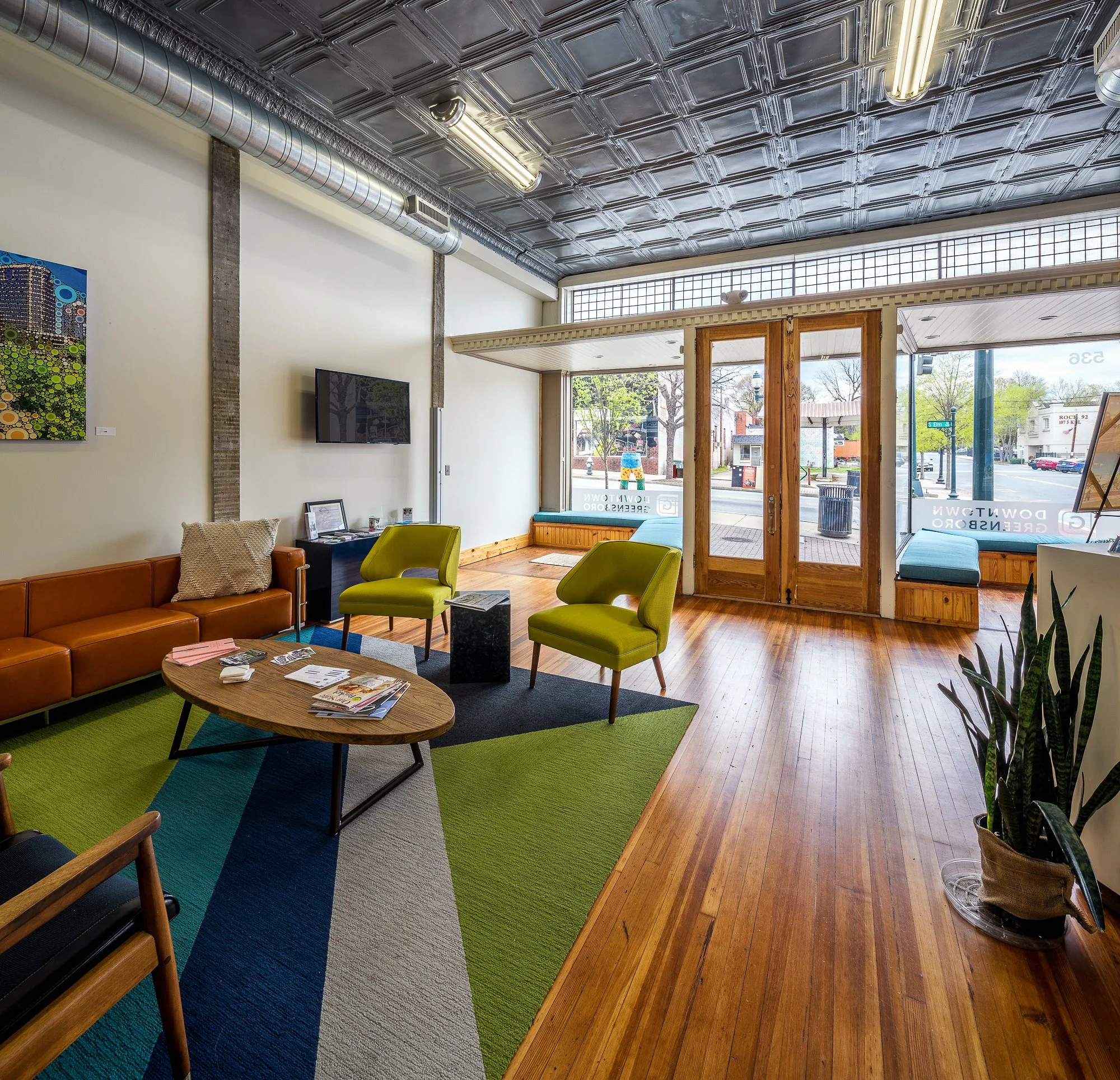 Interior of a cozy waiting or lounge area with a wooden floor, colorful geometric-shaped carpet, bright green chairs, a brown leather couch, and a flat-screen TV on the wall. Large windows with natural light and views of the outside street and trees.